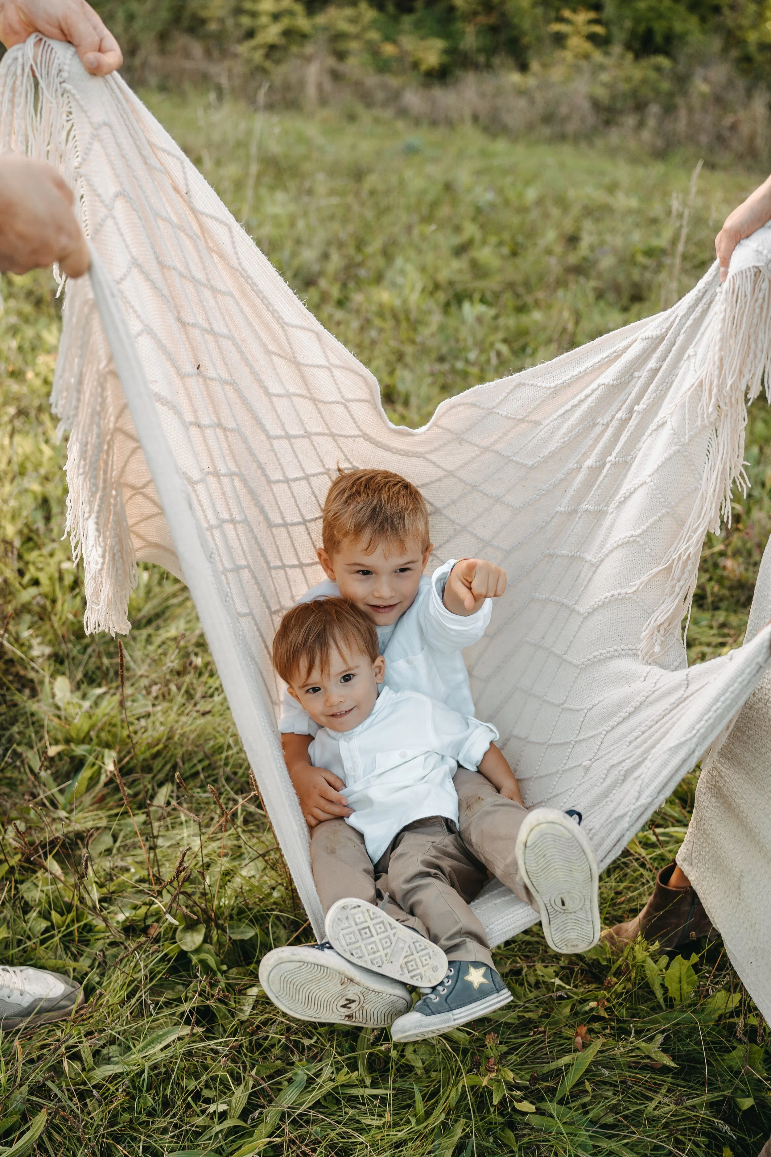 Zwei junge Jungen sitzen in einem weißen Netz in einem Feld, während Erwachsene das Netz halten. Der ältere Junge zeigt mit dem Finger auf etwas außerhalb des Rahmens.