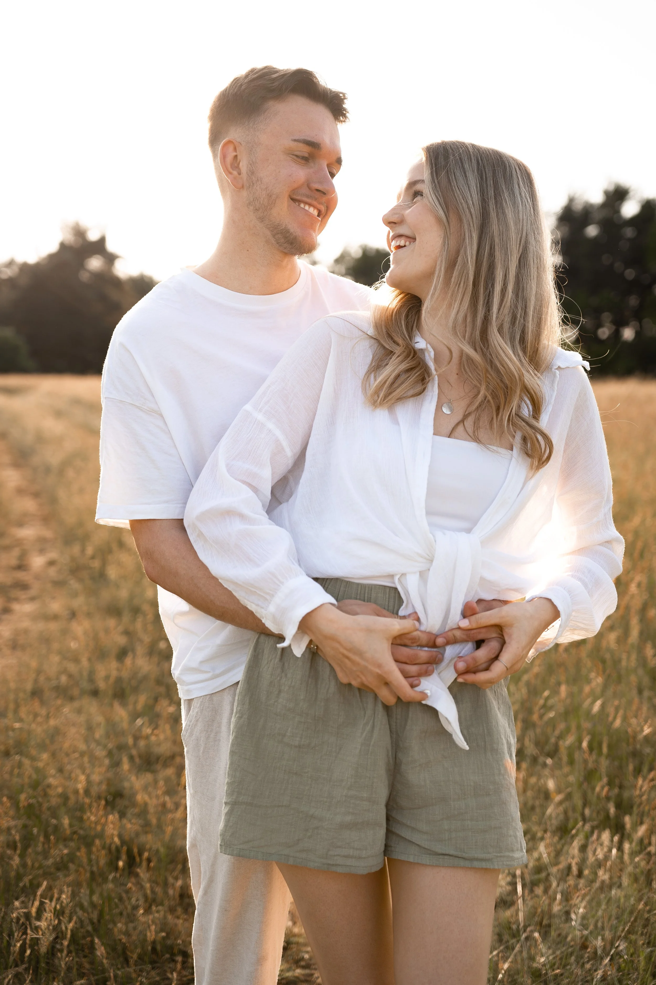 Ein lachendes Paar steht im Sonnenuntergang in einem Feld, umarmt sich liebevoll.