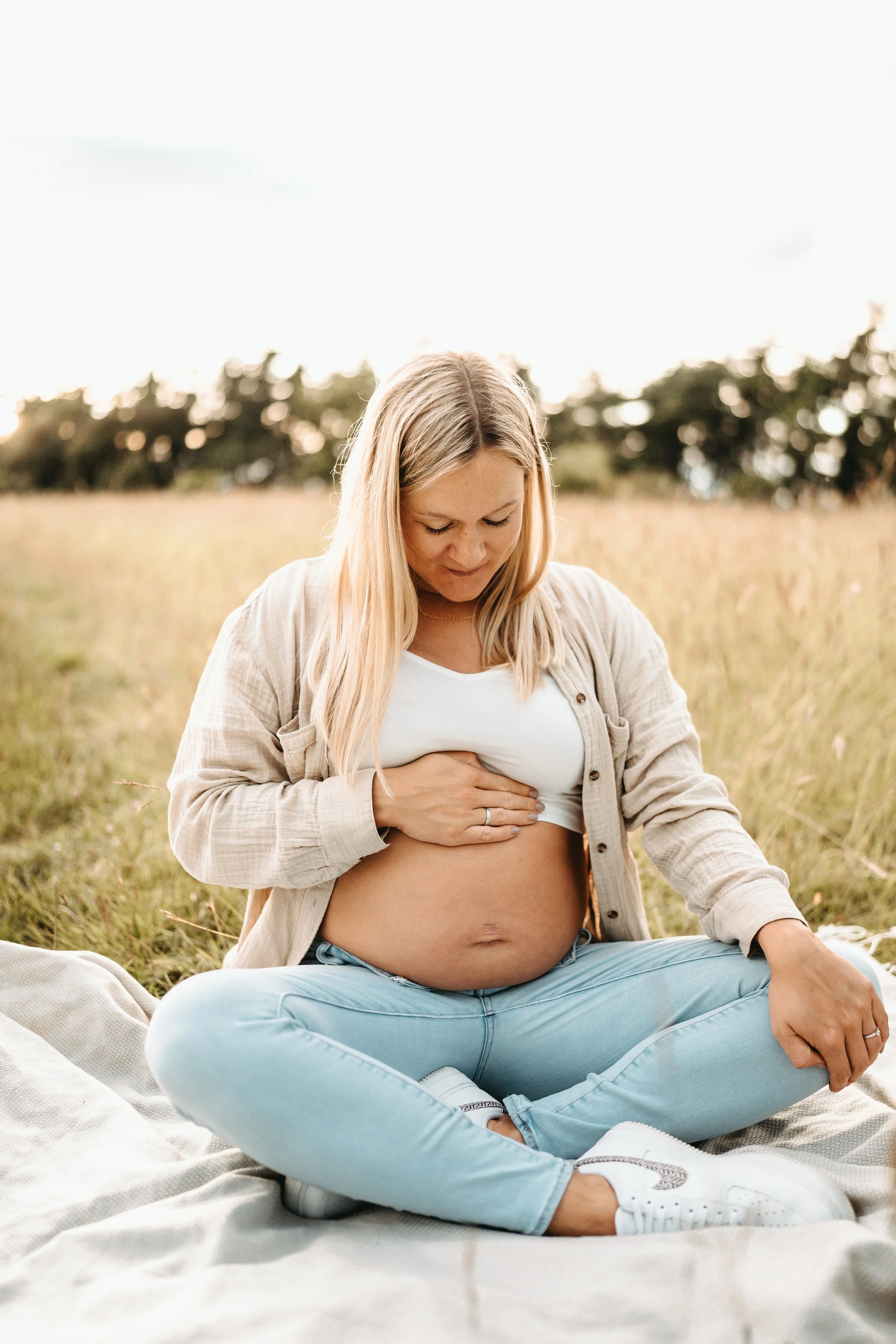 Schwangere Frau sitzt im Freien, hält ihren Bauch, während sie auf einem Bett im Gras sitzt.