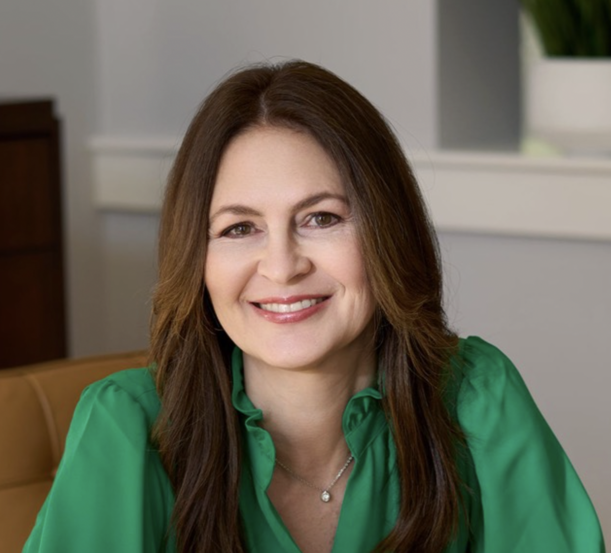 A woman with long brown hair, wearing a green blouse and a necklace, smiling indoors with a window and a houseplant in the background.