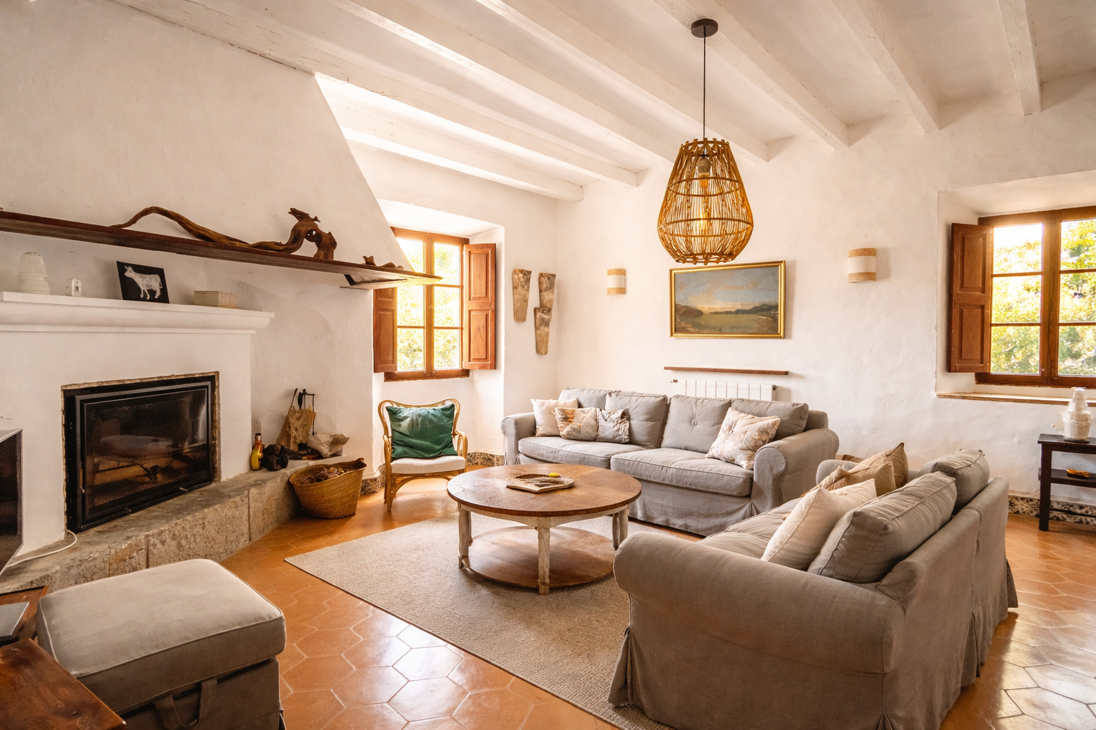 Living room with white stucco walls, wooden beams, a fireplace, a round coffee table, and three couches with pillows. There are windows with wooden shutters, a painting, and a wicker pendant light.