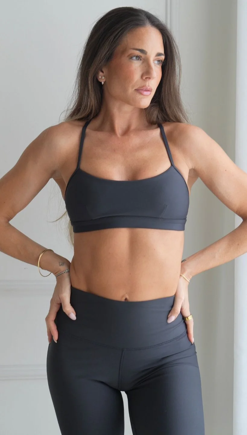 Woman in black sports bra and leggings standing indoors near a window, looking to the side.