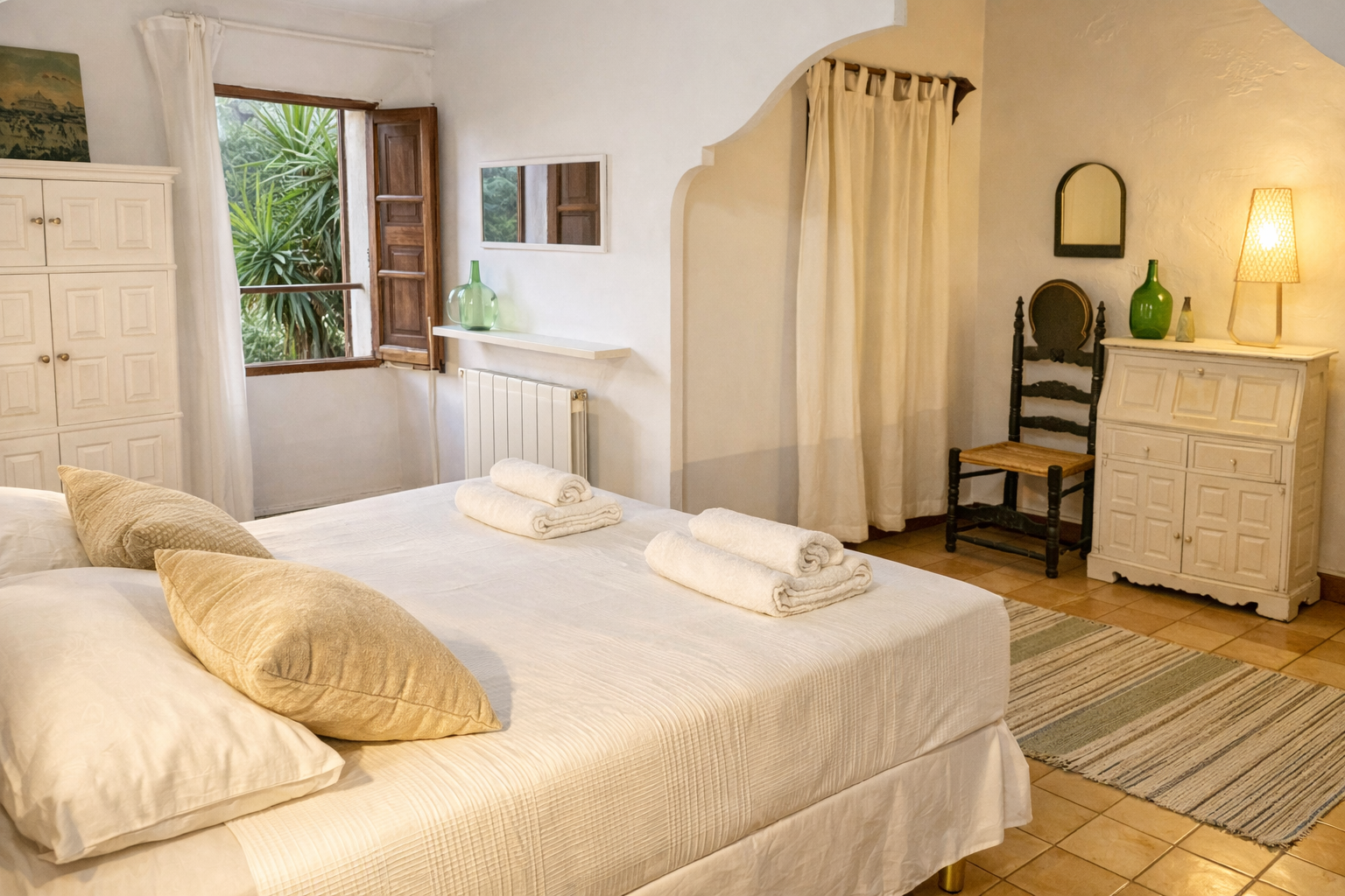 Cozy bedroom with a large bed featuring white linens and beige pillows, open window with wooden shutters revealing green foliage outside, white furniture including a dresser with a green vase and a reading lamp, a black chair, and a striped rug on a tiled floor.
