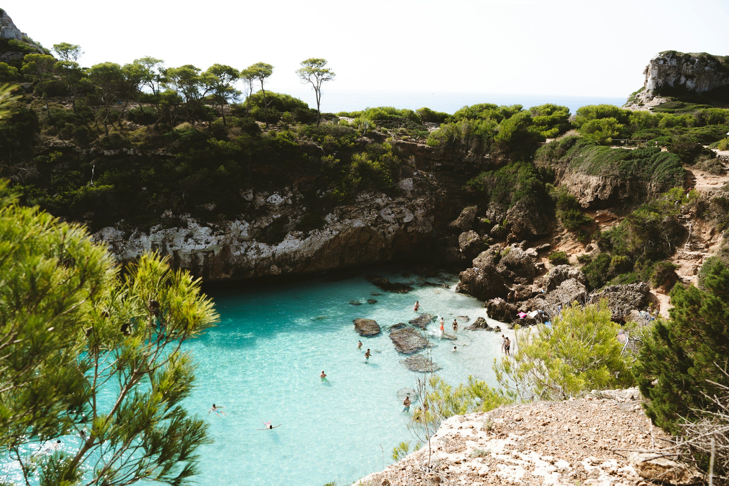 A secluded beach with turquoise water, surrounded by rocky cliffs and lush green trees, with people swimming and relaxing in the water.