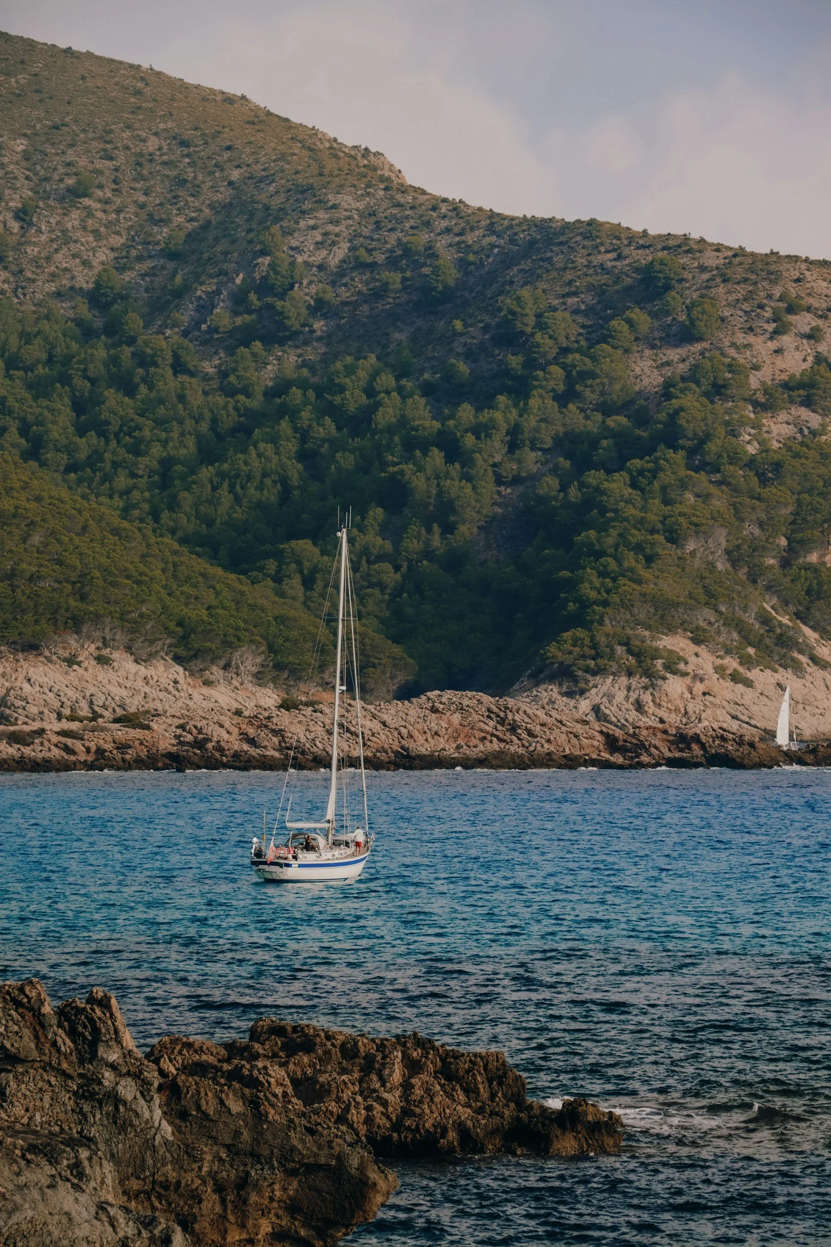 A sailboat on calm blue waters near a rocky shoreline with green hills and a partly cloudy sky in the background.