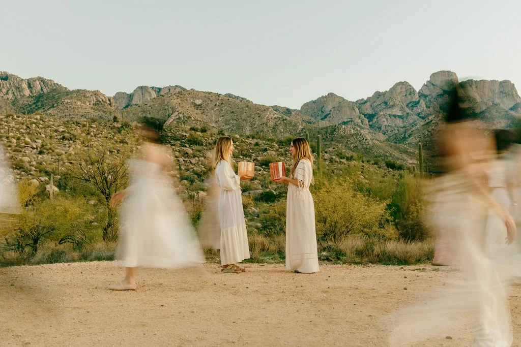 Two women in white dresses face each other while holding books or jars, with a desert landscape and mountains in the background. Several blurred figures in white dresses walk by in the foreground.