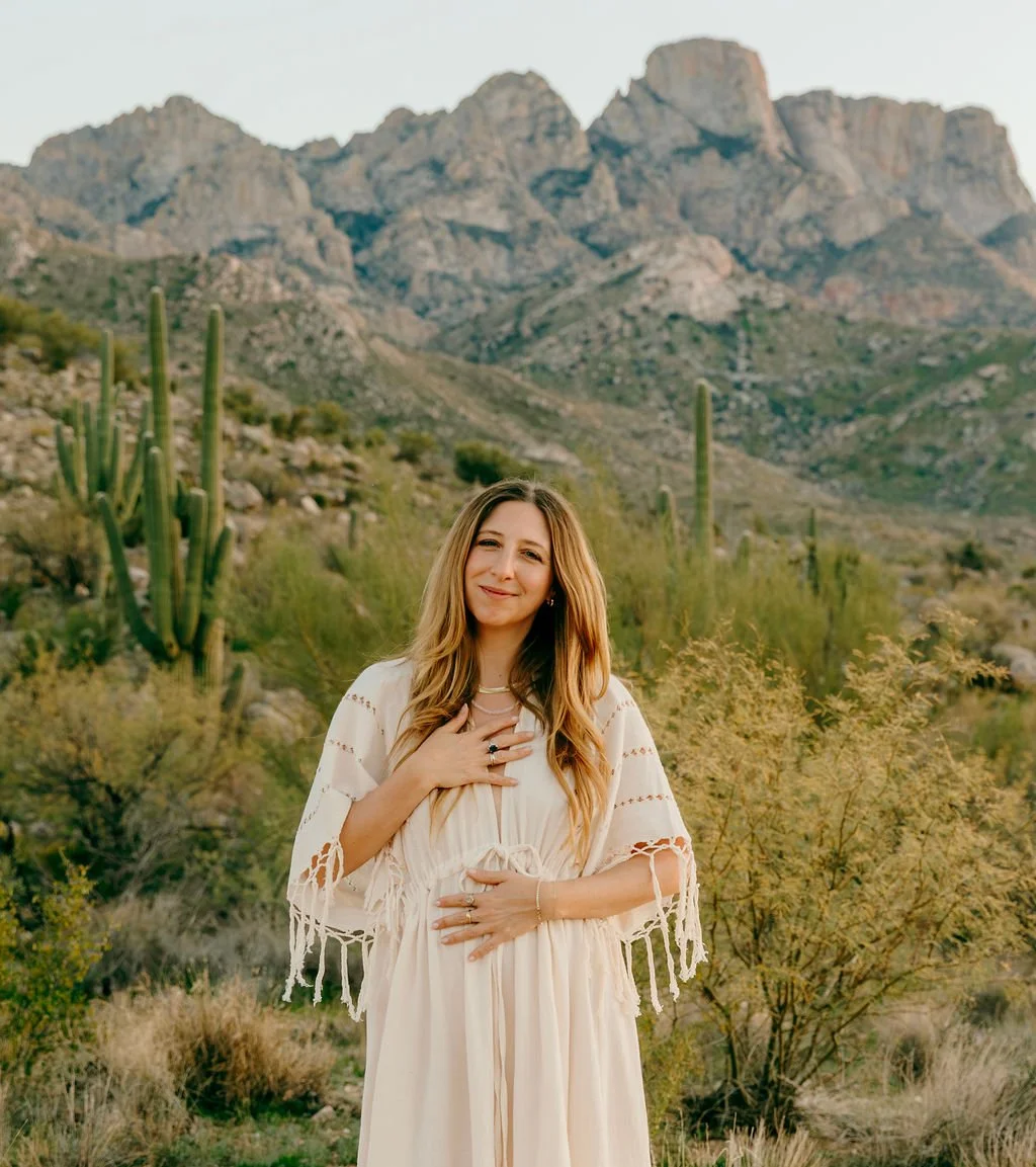 A young woman with long blonde hair wearing a white dress and shawl, standing in a desert landscape with cacti and mountains in the background.