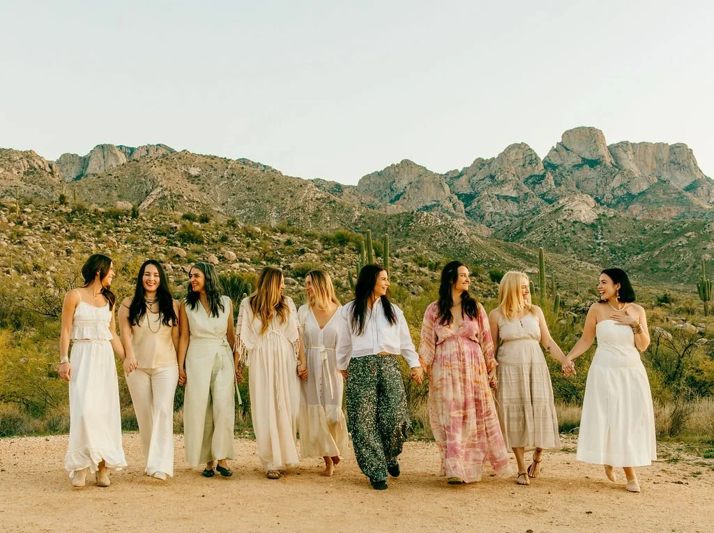 Group of women holding hands and walking outdoors in a desert landscape with mountains and cacti in the background.