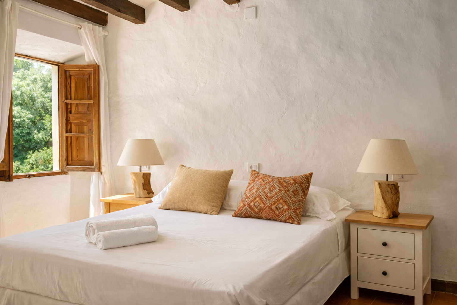 A cozy bedroom with white textured walls, a window with wooden shutters, a bed with white linen, two beige and patterned decorative pillows, and two matching bedside tables with lamps made from wooden bases and cream lampshades.
