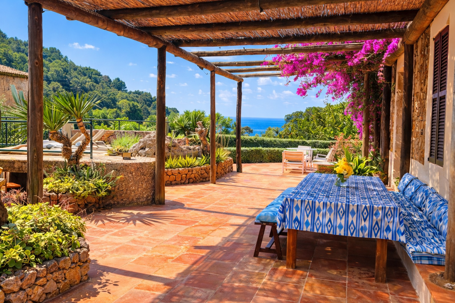 A terracotta tiled outdoor patio with a blue patterned tablecloth on a wooden table, surrounded by greenery, flowering plants, and a view of the ocean in the distance.