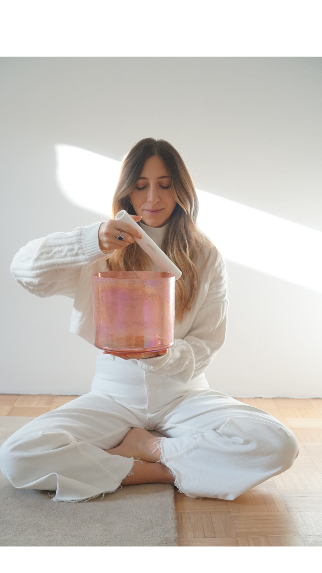 A woman sitting cross-legged on a wooden floor, holding a pink candle holder, and pouring wax into it from a white container.