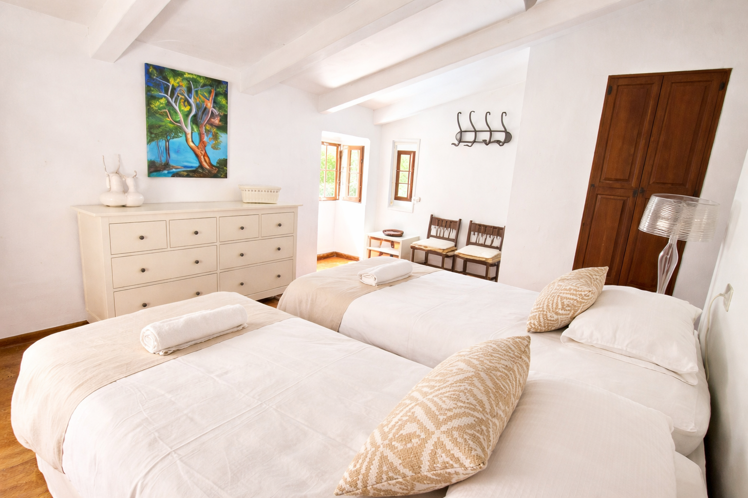 A bright bedroom with two twin beds, white bedding, and rolled towels. The room has a white dresser, a colorful painting of a tree, wooden chairs, and a wardrobe. The walls are white, and there are small windows letting in natural light.