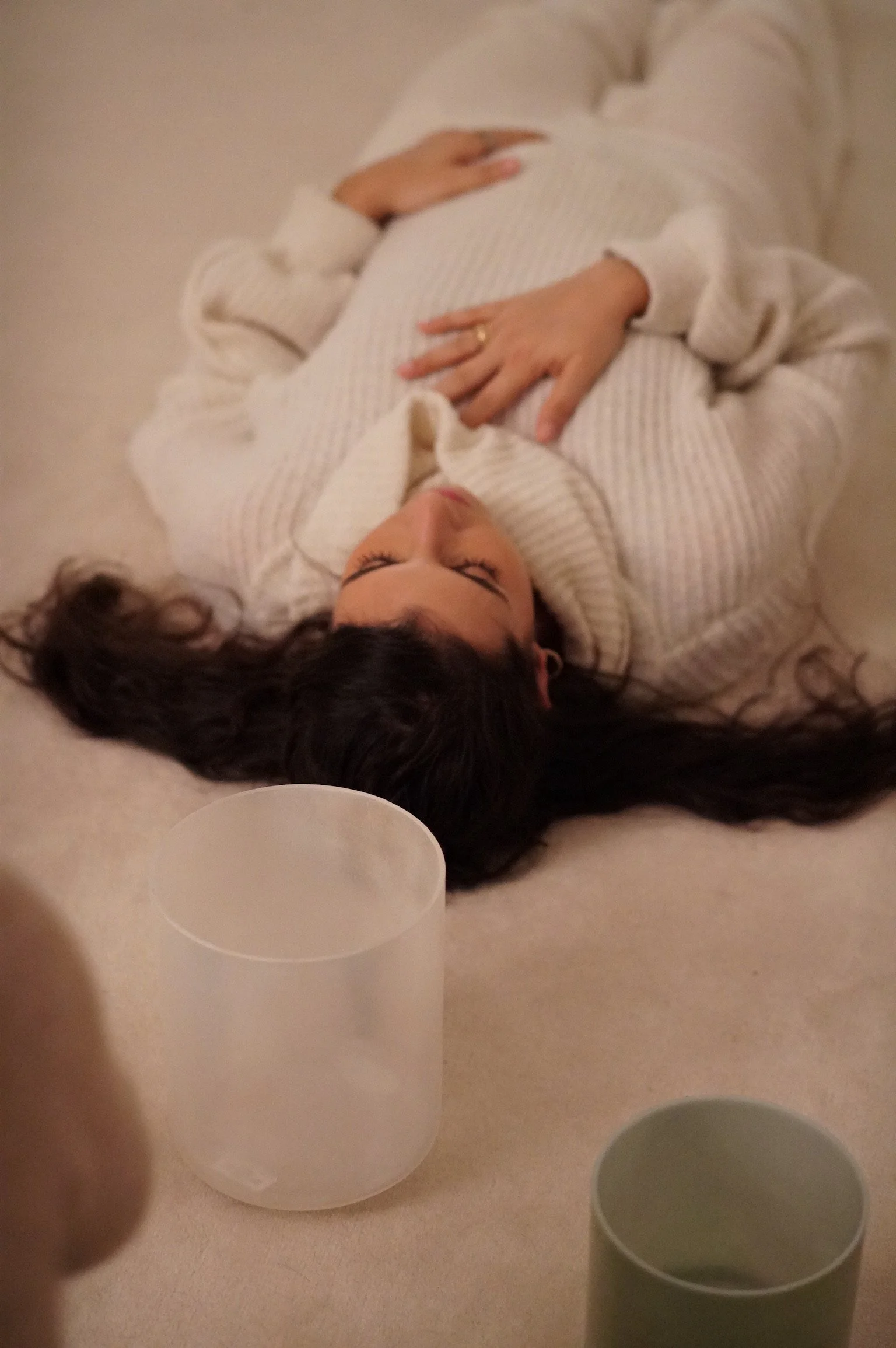 A woman lying on her back on a soft surface, with her eyes closed, hands resting on her pregnant belly, wearing a white sweater, surrounded by two cups or containers.