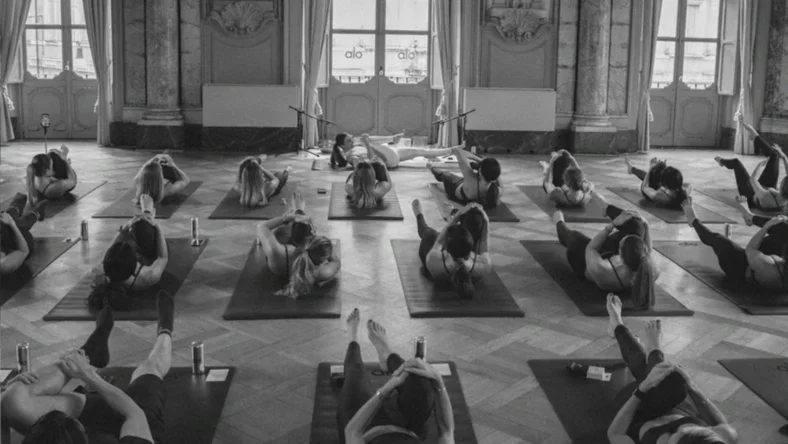 Group of people practicing yoga in a spacious, ornate room with large windows and classical decor, all on yoga mats.