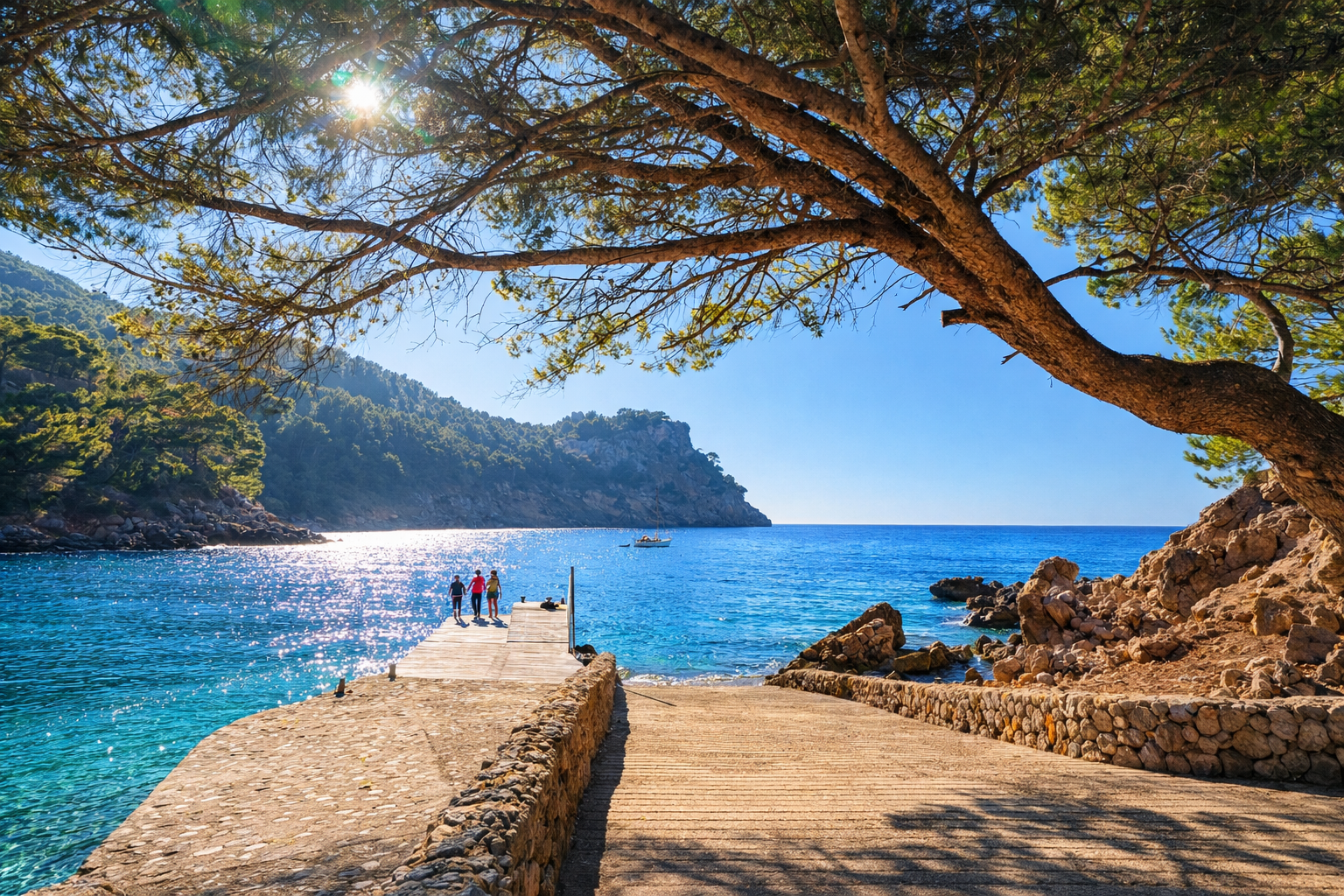 A scenic coastal view with a stone pathway leading to a dock, three people walking on the dock, a sailboat on calm blue waters, rocky shoreline, green hillside, and a large tree providing shade in the foreground, under a clear sky.