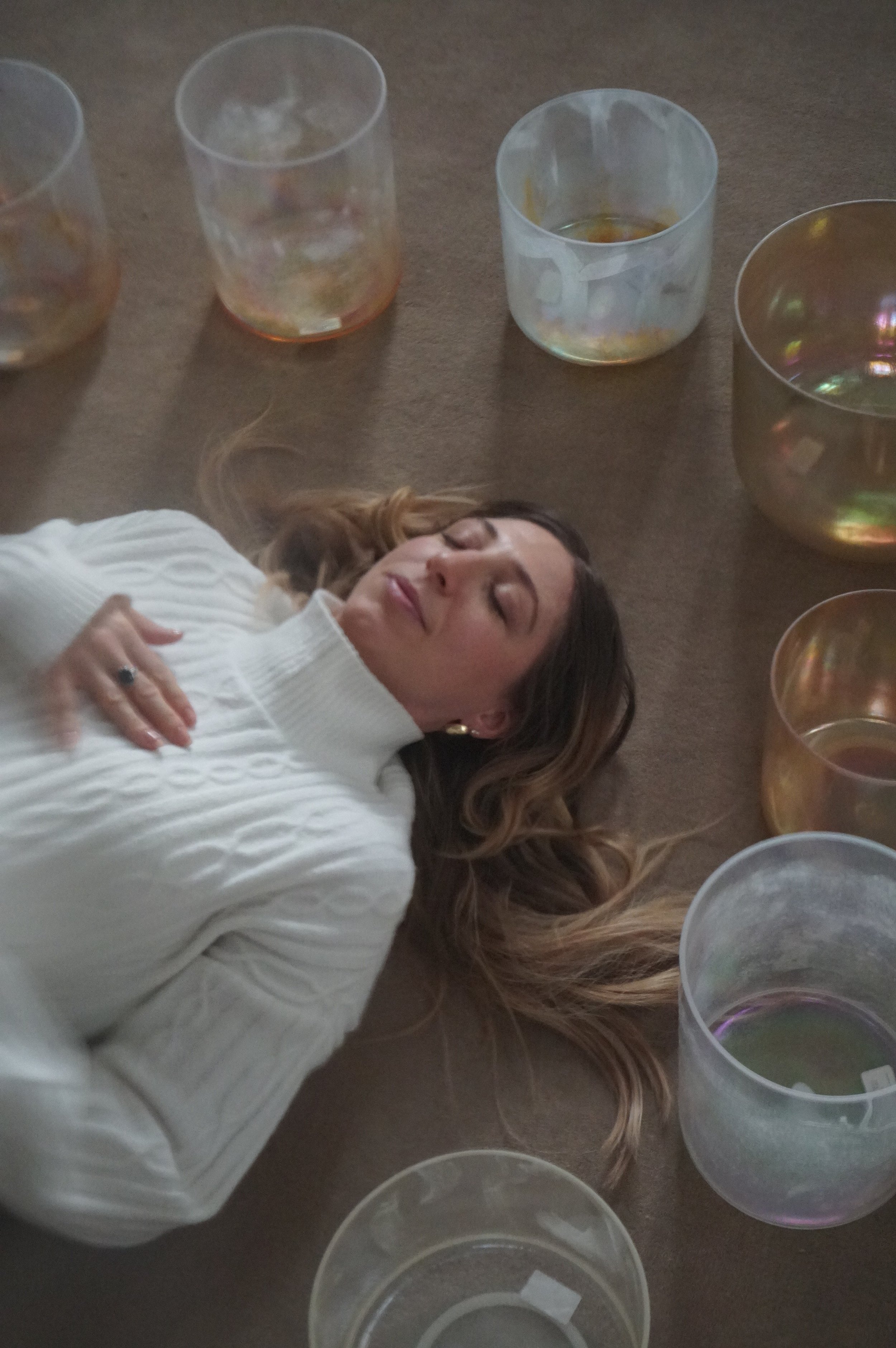 A woman lying on a brown carpeted floor surrounded by various clear and iridescent crystal bowls, wearing a white sweater with her eyes closed.