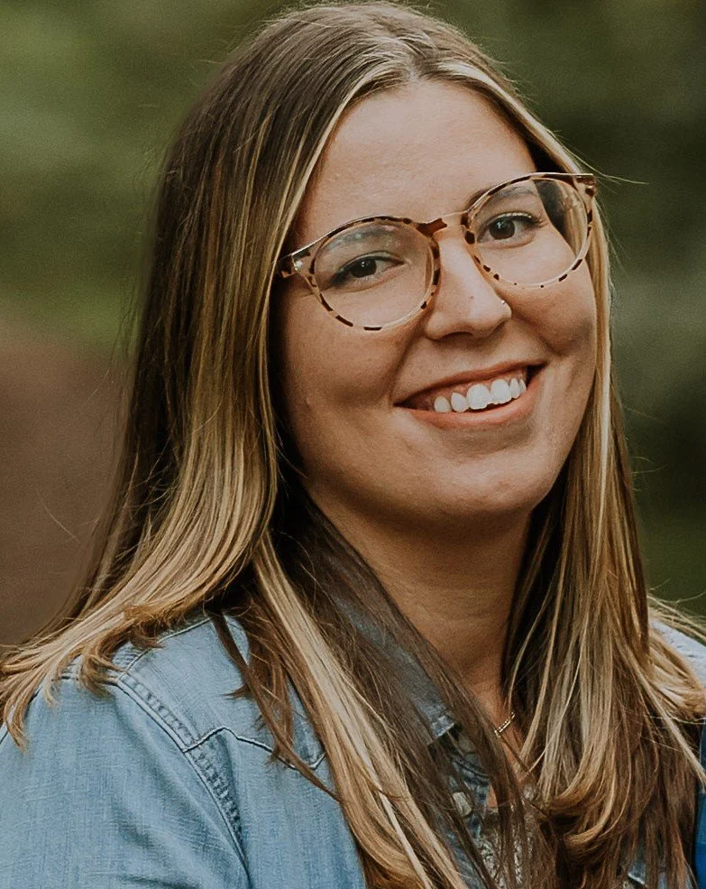 A young woman with long blonde hair, wearing glasses and a denim jacket, smiling outdoors.