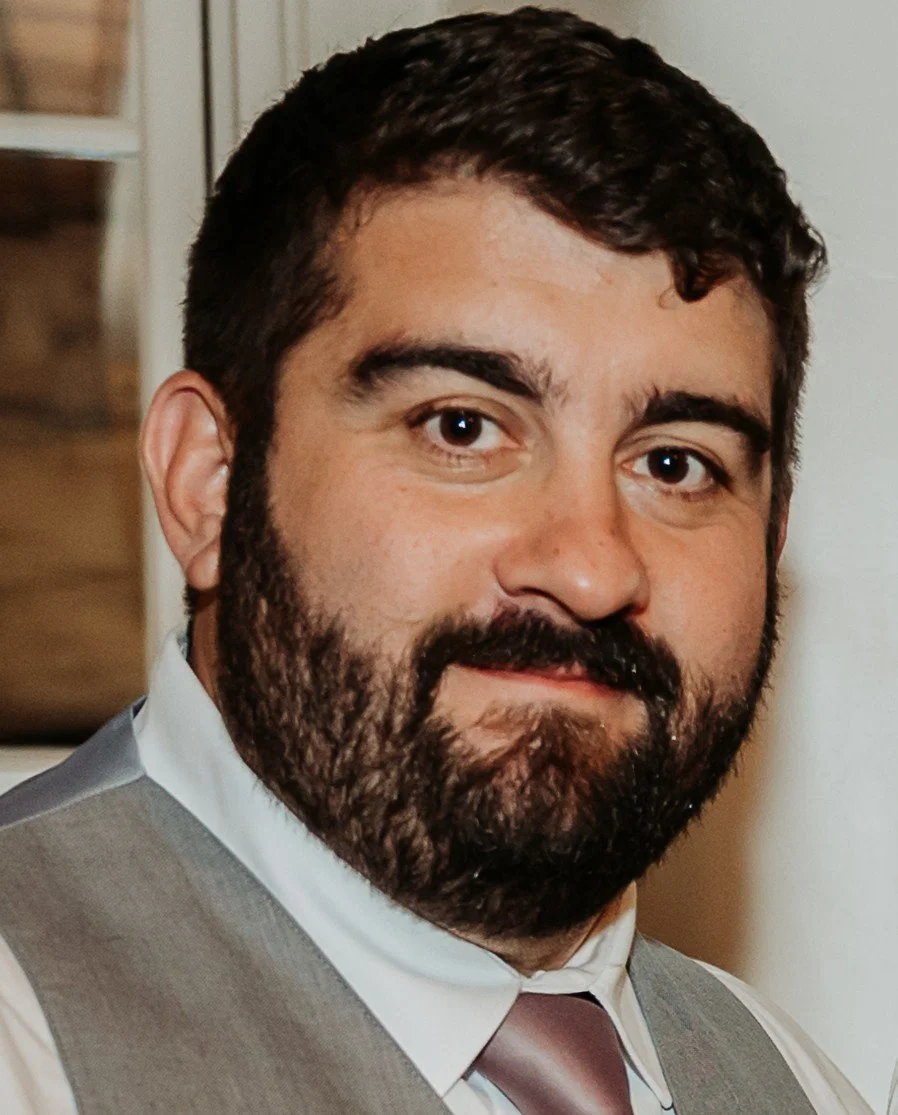 A man with dark hair and a beard, wearing a gray vest, white shirt, and a muted pink tie, looking at the camera with a slight smile.