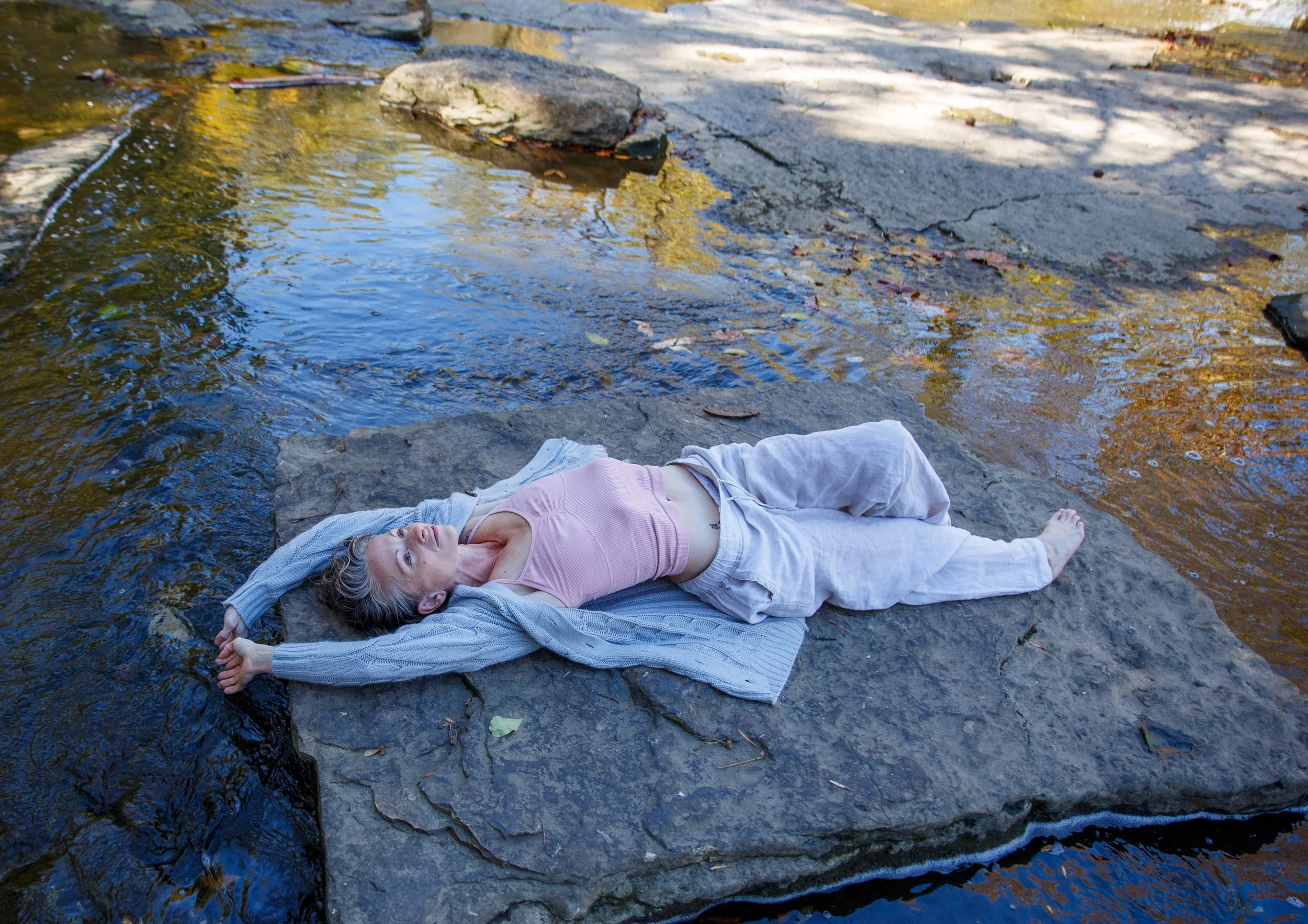 Woman lying on a large flat rock by a river, relaxing with her arms behind her head, wearing a pink top, gray pants, and a gray cardigan.