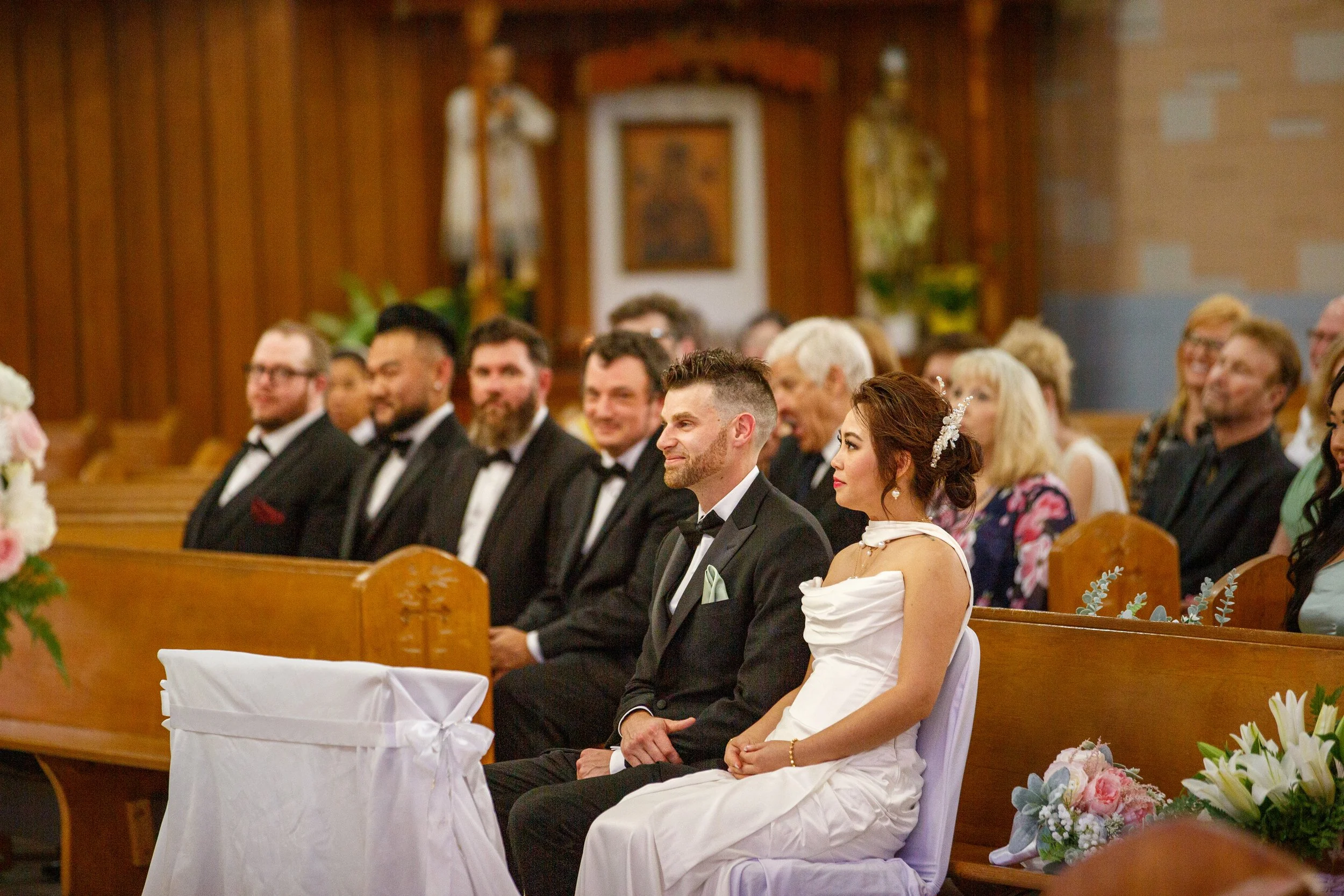 Bride and groom sitting in a church pew during their wedding ceremony, surrounded by guests, with the bride wearing a white dress and the groom in a black tuxedo.
