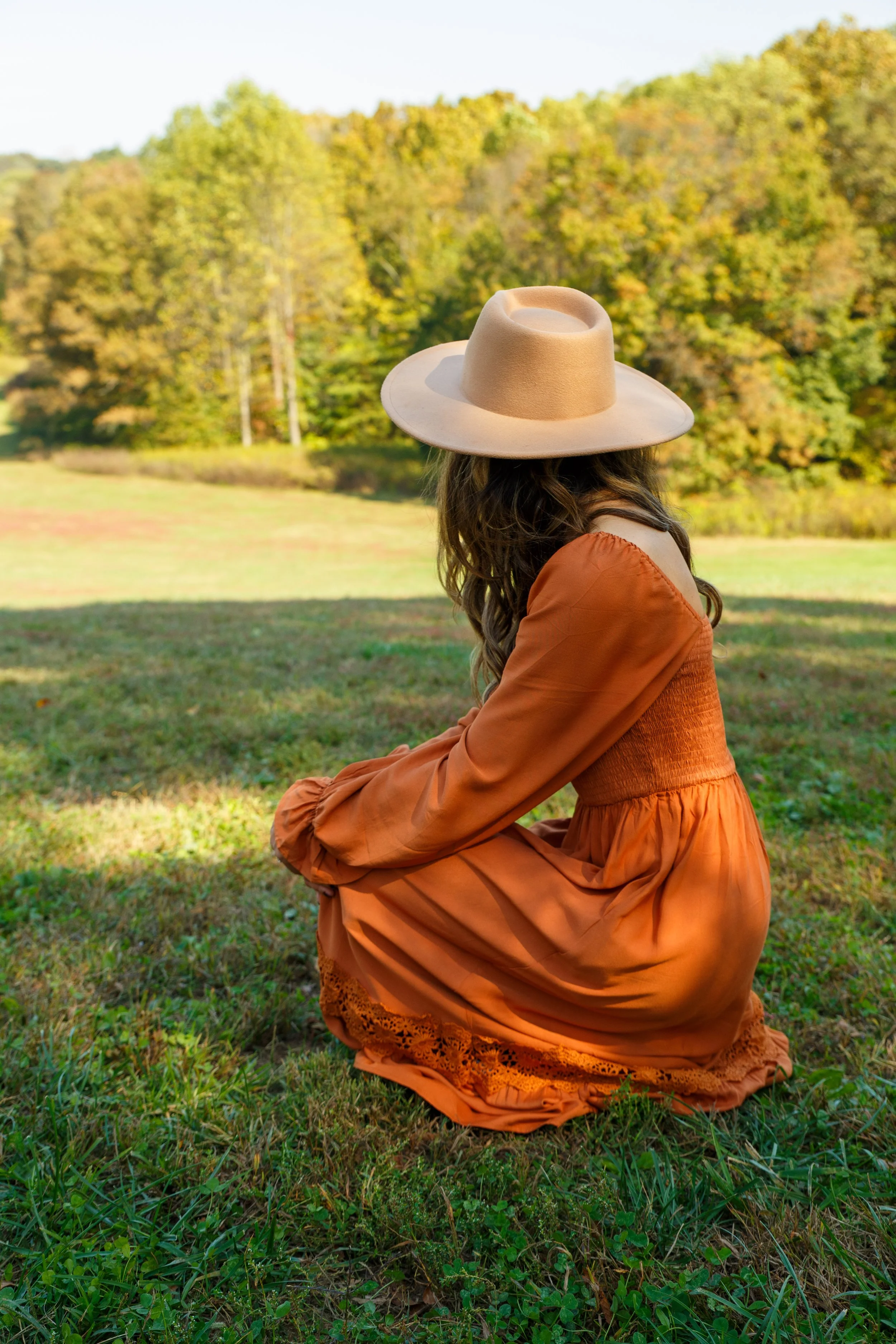 Woman wearing an orange dress and wide-brimmed beige hat, squatting in a grassy field with trees in fall colors in the background.