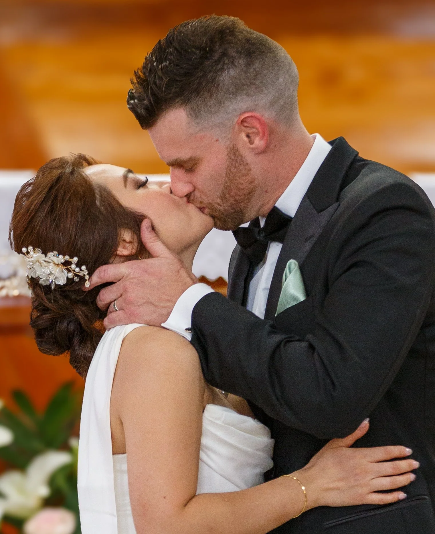 A bride and groom sharing a kiss at their wedding ceremony, with the groom holding the bride's face and the bride holding the groom's face, in an indoor setting with warm lighting.