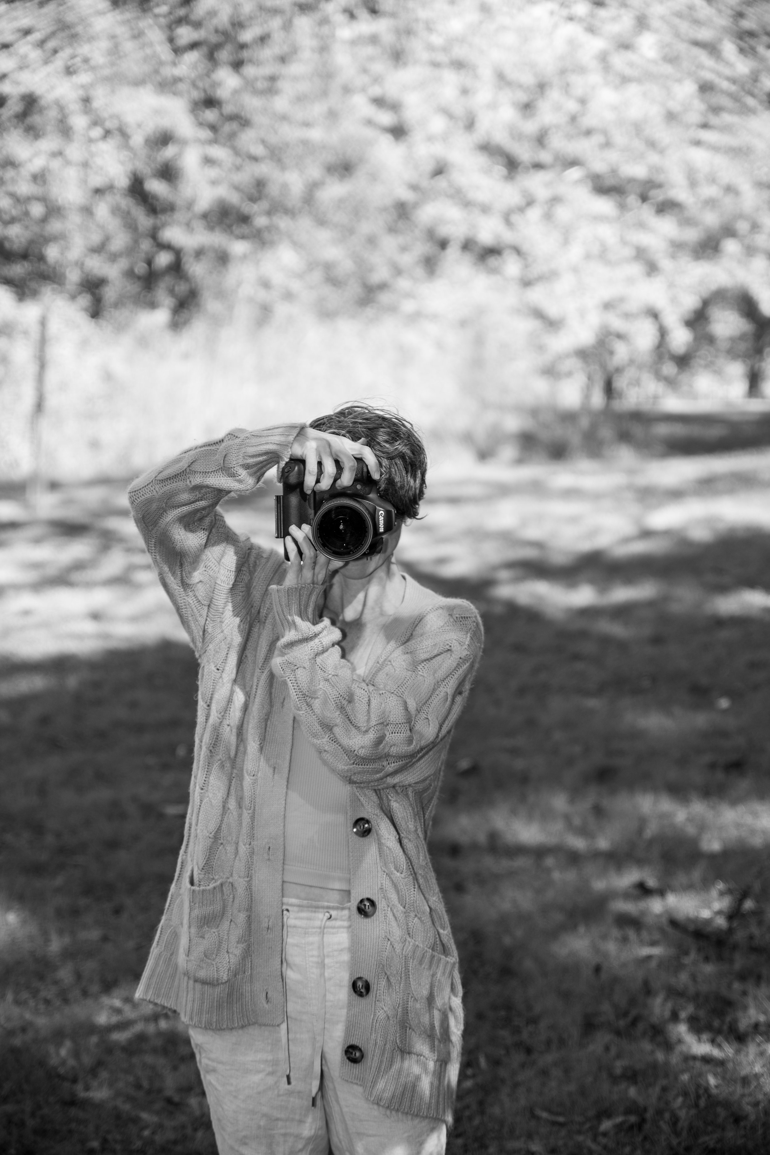 A person standing outdoors in a park, holding a camera up to their face, taking a photograph. The person is wearing a cable knit cardigan and light-colored pants. Trees with autumn foliage are visible in the background.