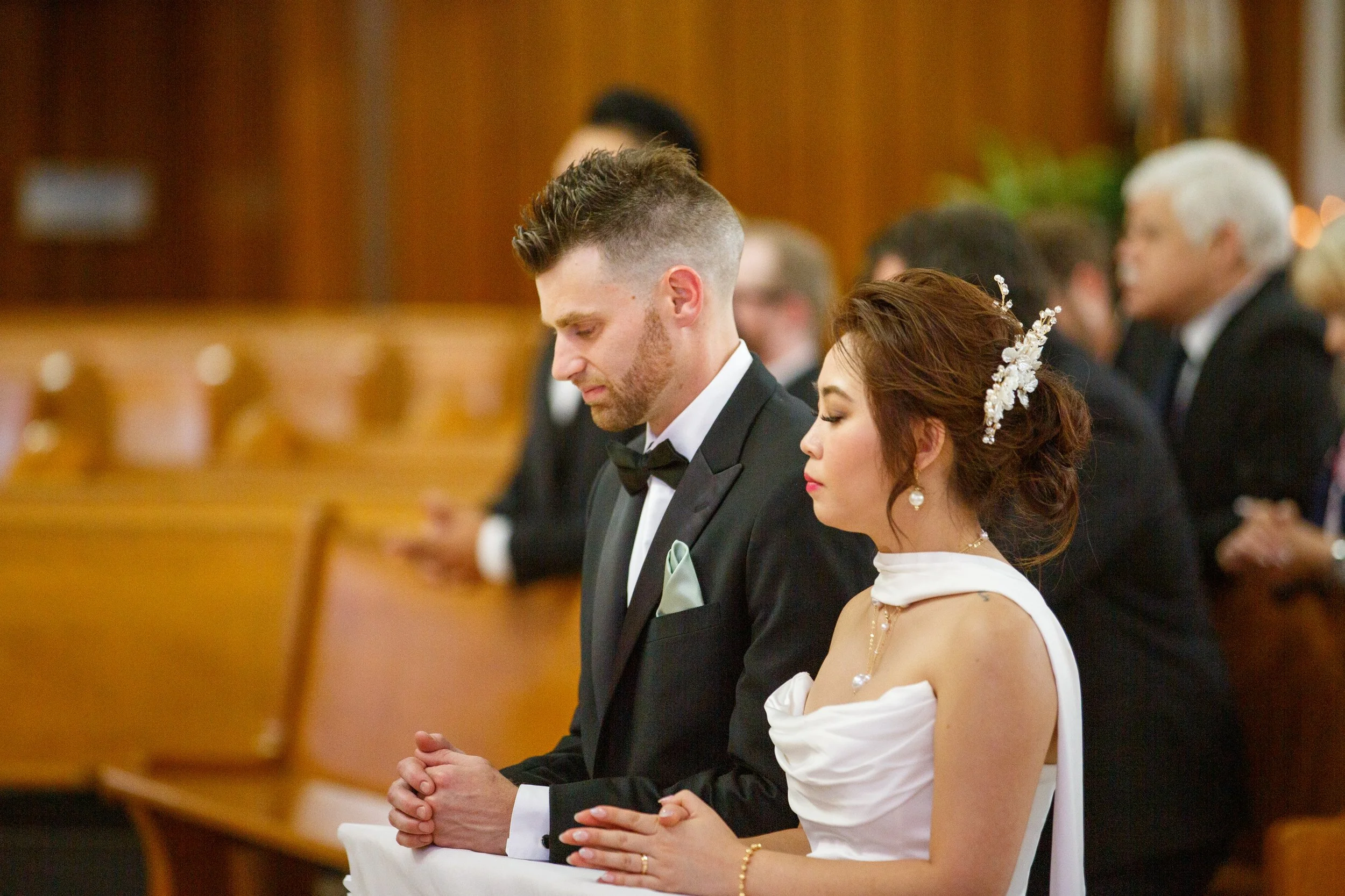 A bride and groom kneeling in prayer during a wedding ceremony in a church, with their eyes closed and hands clasped.