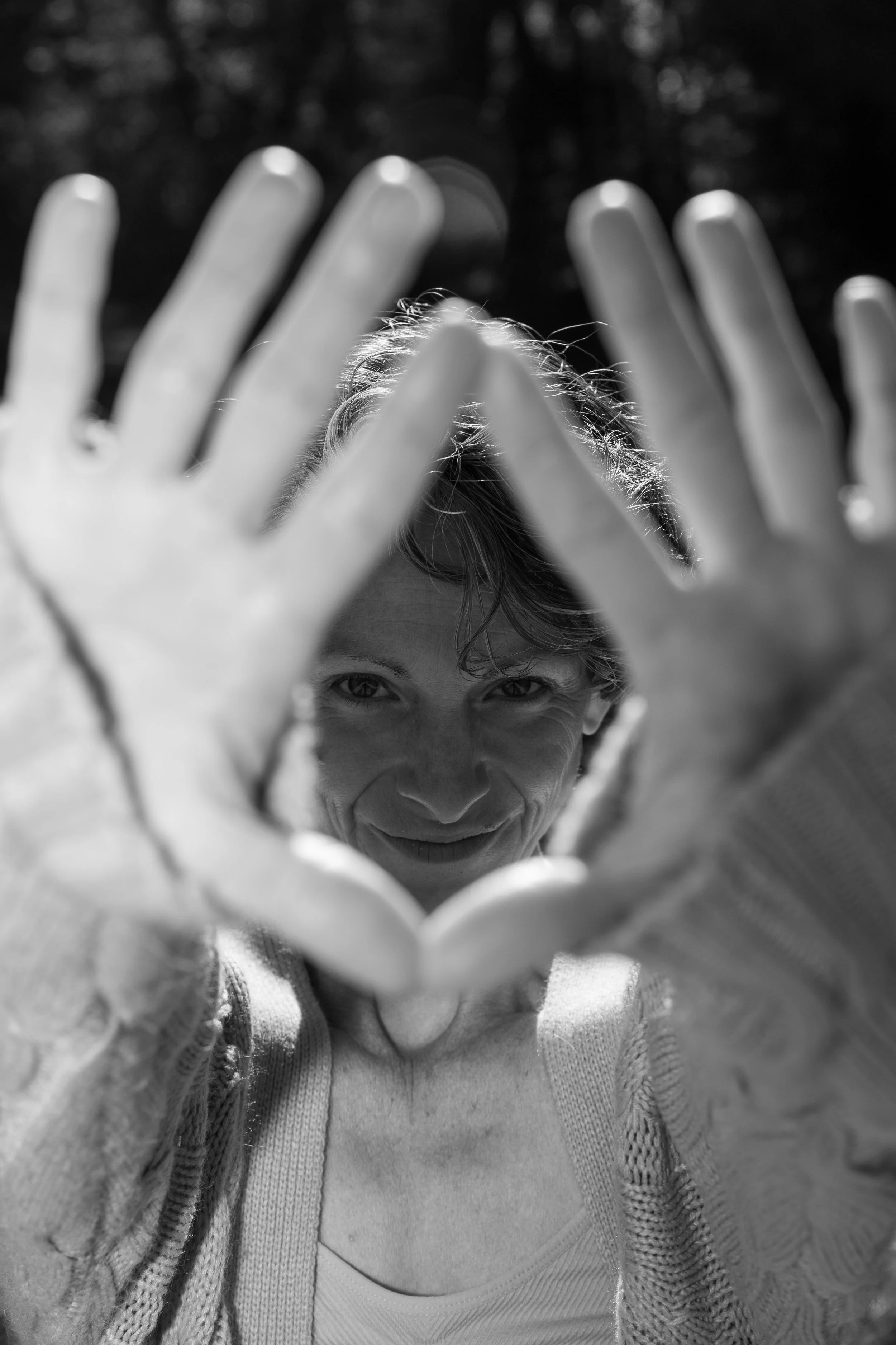 A woman smiling through her hands, which are held up in front of her in a heart shape, outdoors in sunlight.