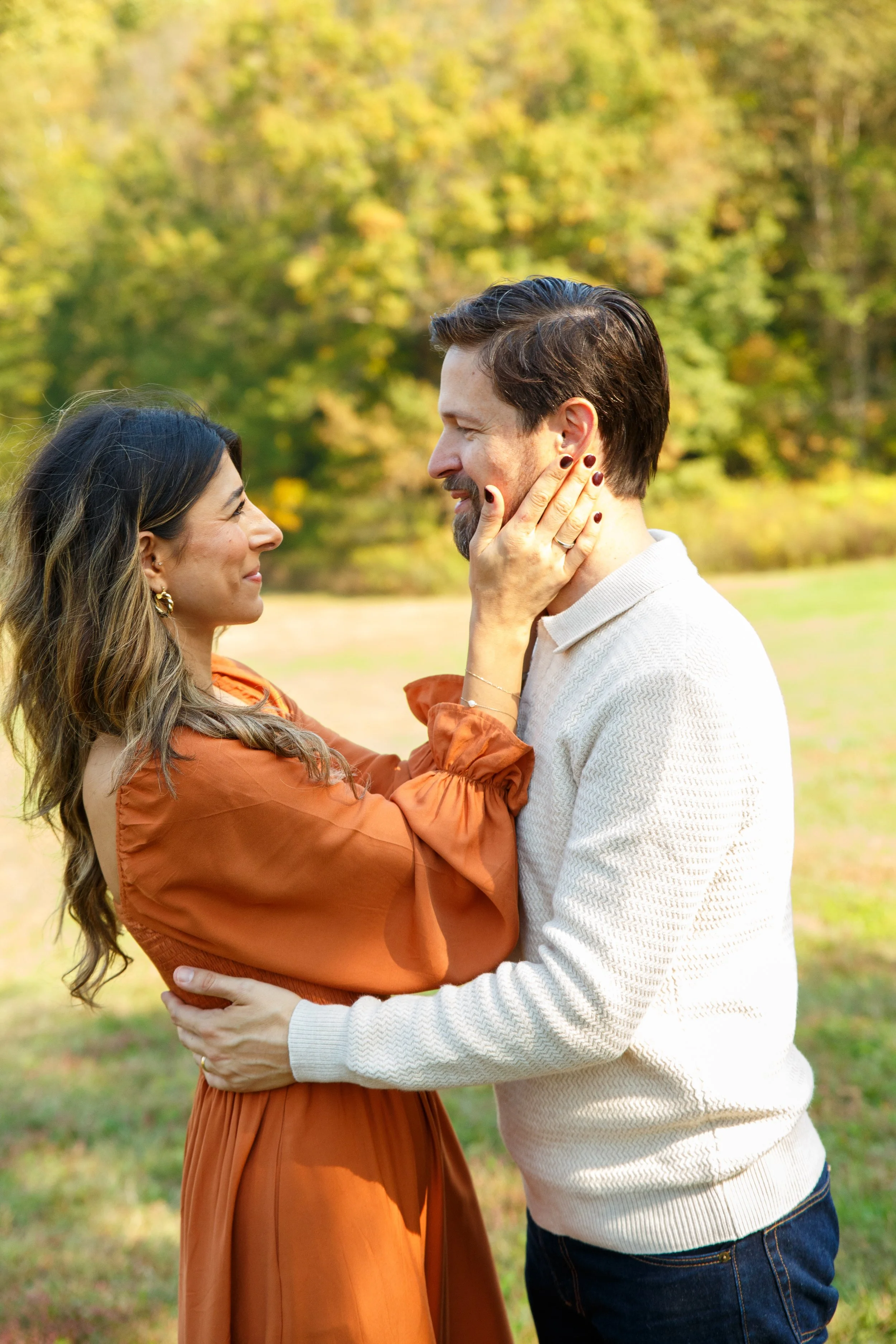 A couple standing outdoors in a park, embracing and looking into each other's eyes with autumn trees in the background.