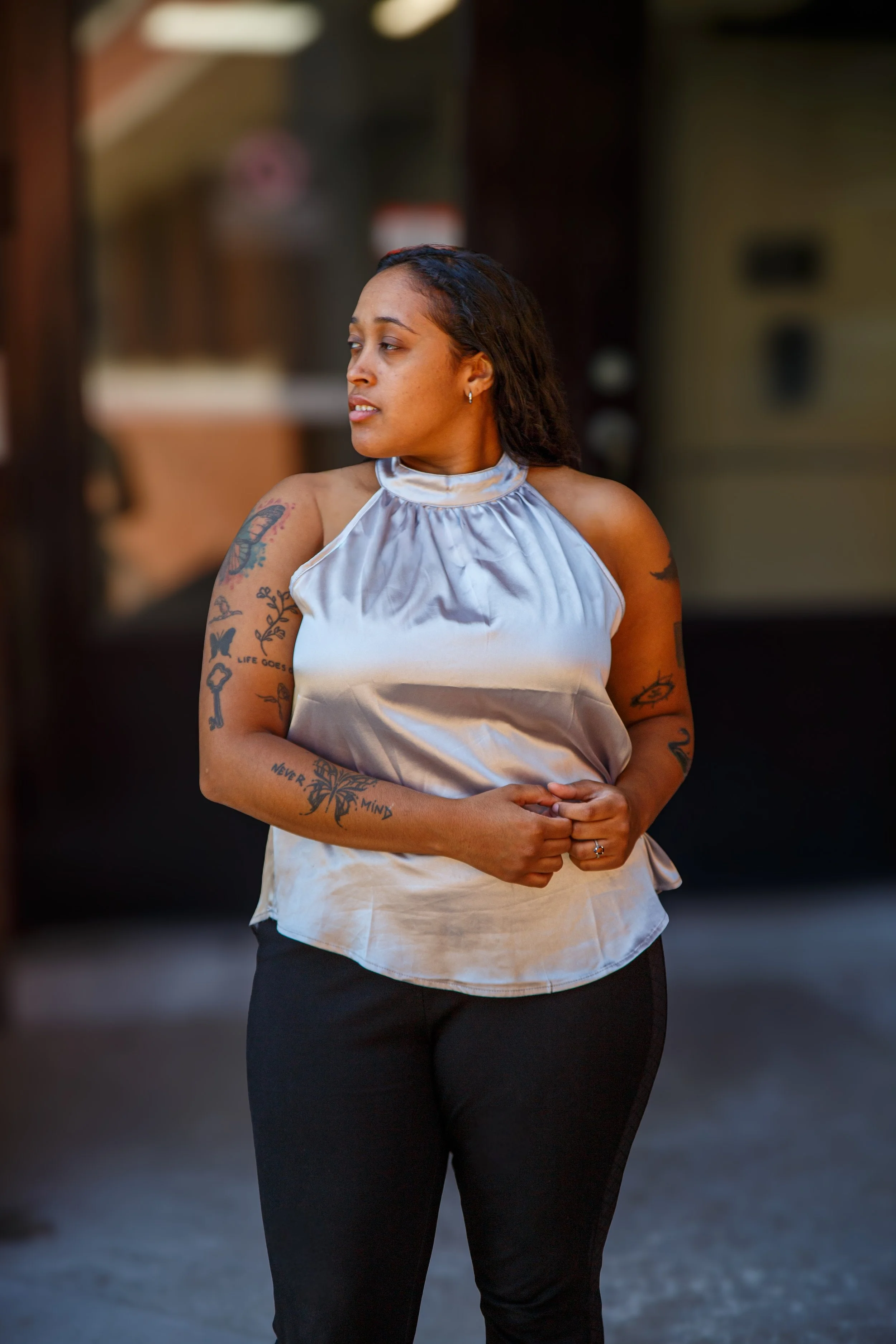 A woman with tattoos on her arms standing outdoors, looking to her left, wearing a shiny silver sleeveless top and black pants.