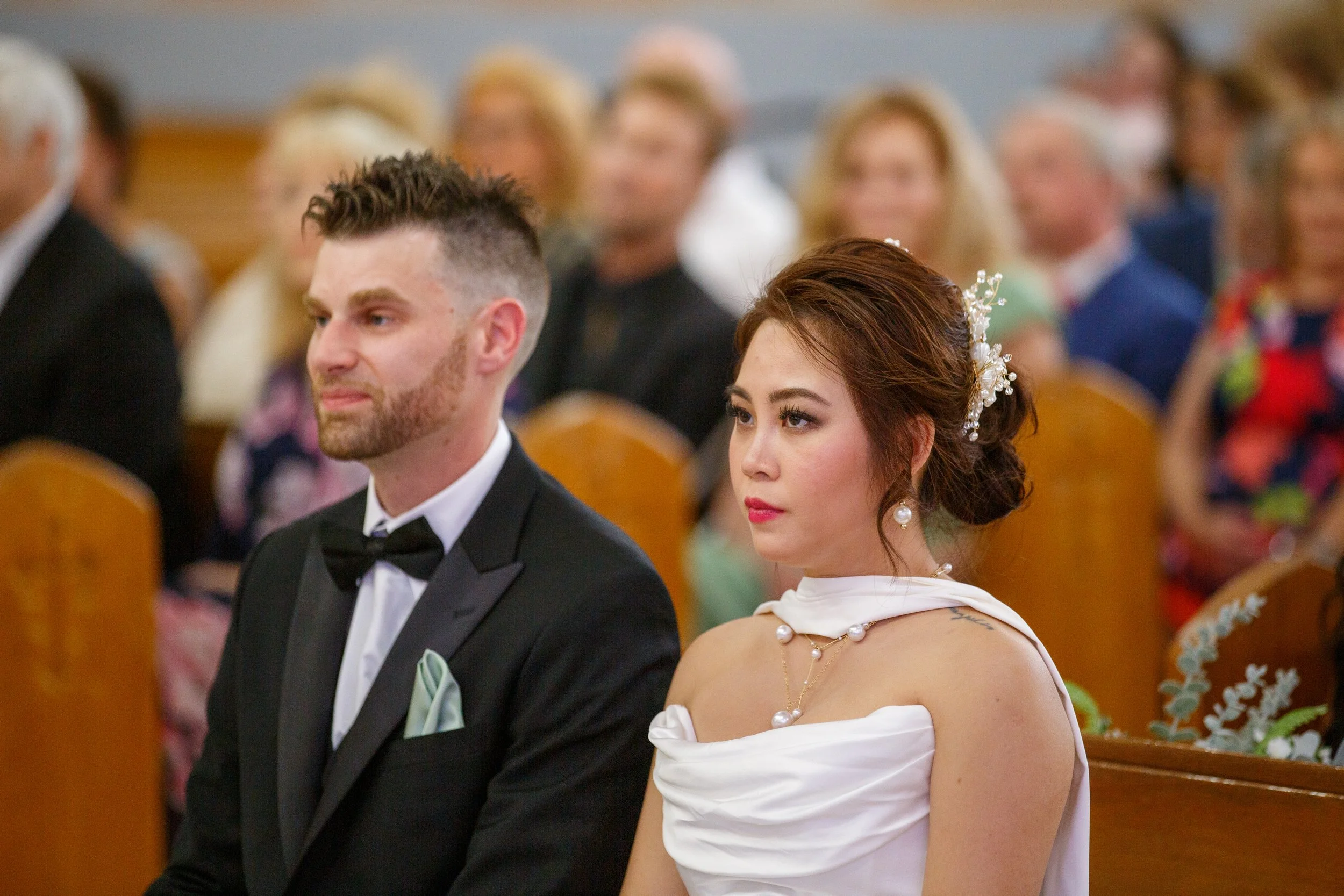 A bride and groom sitting during a wedding ceremony, with the bride in a white strapless gown and the groom in a black tuxedo with a bow tie, surrounded by guests seated in wooden church pews.