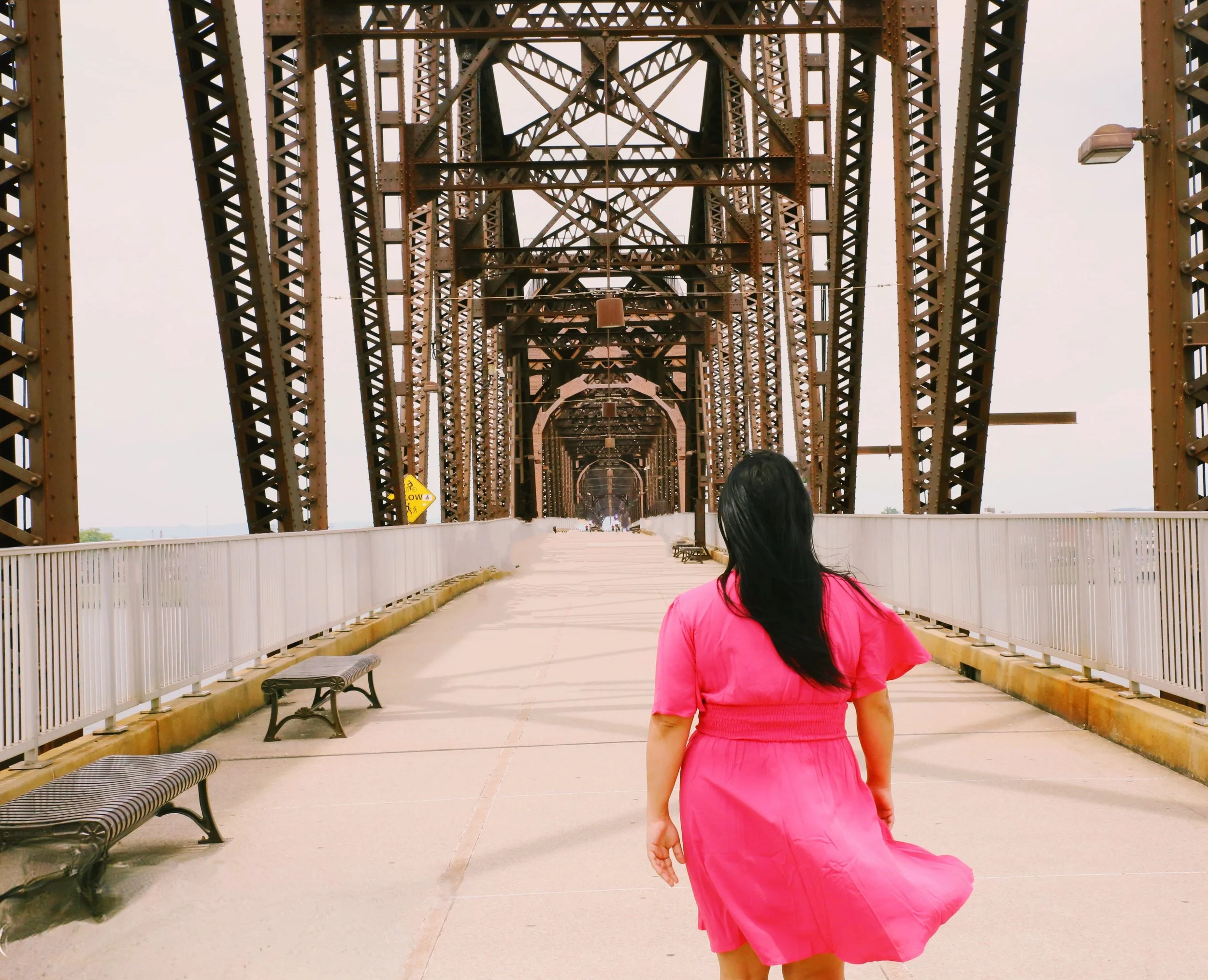 A woman with long black hair wearing a pink dress walking along a pedestrian bridge with a metal truss structure, benches on the side, and a cloudy sky.