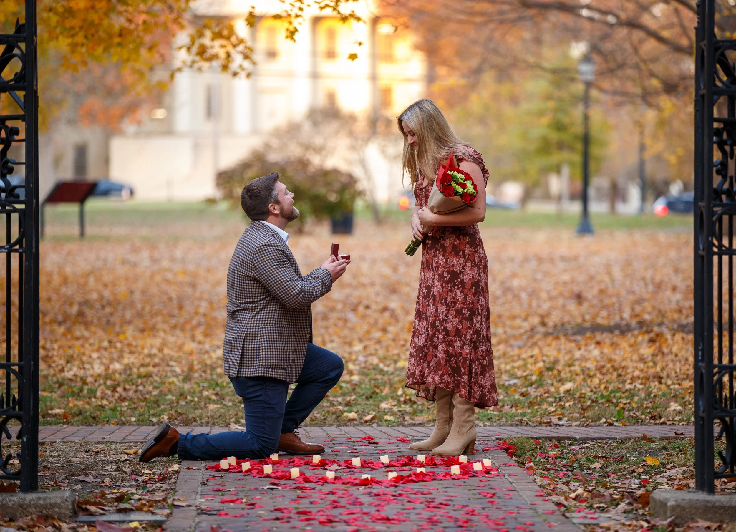 A man proposing to a woman on a fall day in a park, with pumpkin candles and rose petals on the ground, a woman holding a bouquet of flowers, and man on one knee.