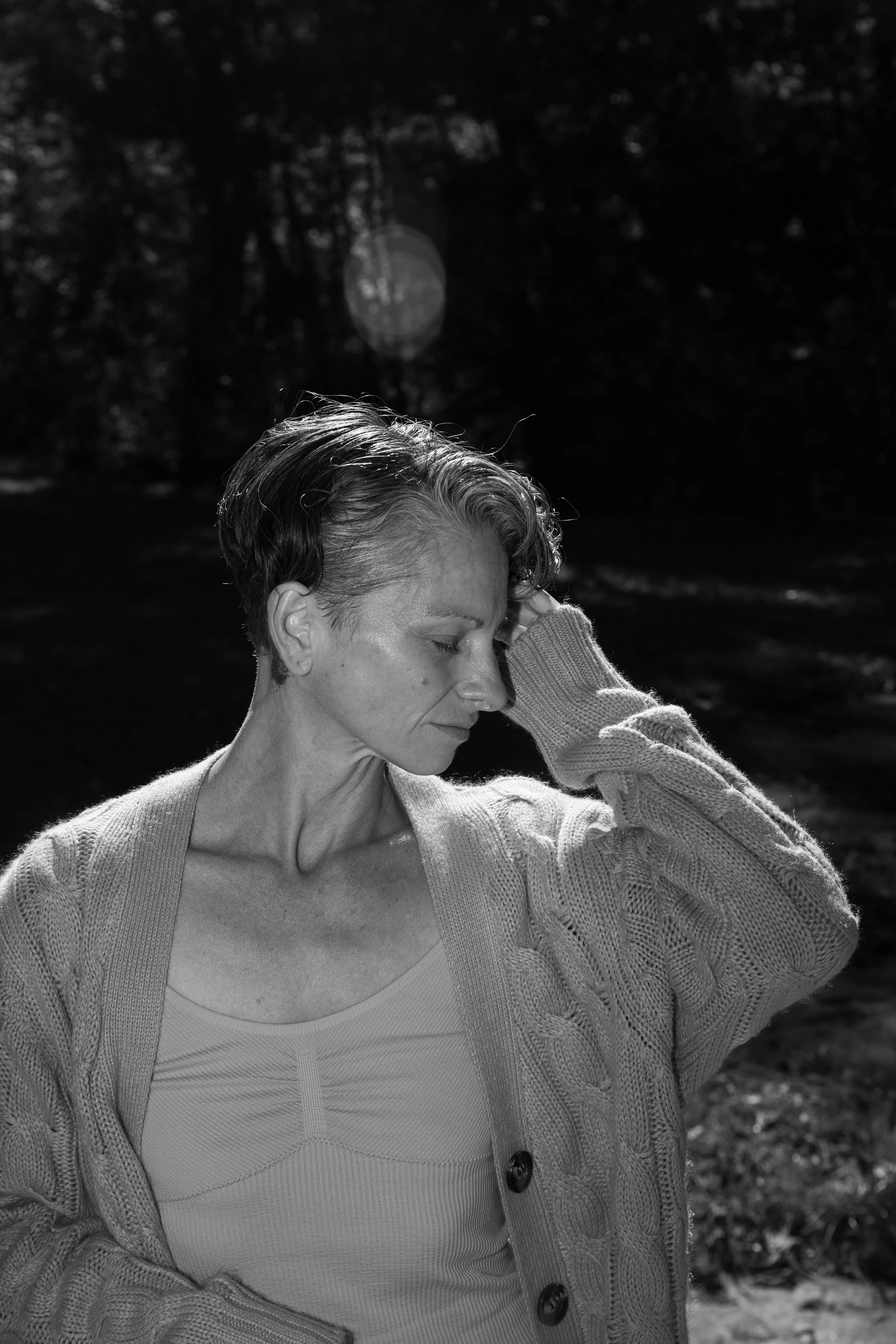 A black and white photo of a woman with short hair, wearing a knit sweater and tank top, standing outdoors with her eyes closed and hand resting on her head.