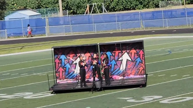 Four people performing on a small stage set up on a football field, with a colorful graffiti-style backdrop featuring arrows.