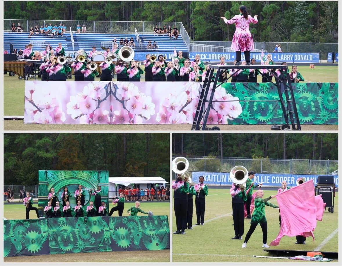 A marching band performing on a sports field, with a conductor on a raised platform. The band members are wearing green and pink uniforms, and some are playing brass instruments. Behind them, there are spectators seated in bleachers, and banners with names of schools or sponsors are visible. The scene appears to be during a halftime show or a similar event.