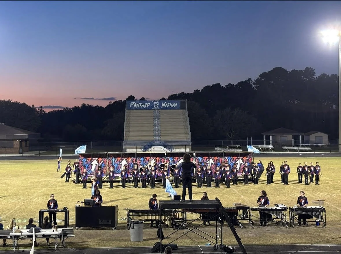 A school marching band performs on a football field at sunset. The band members wear purple and black uniforms, and large banners with colorful arrows are displayed behind them. There are empty bleachers in the background with a sign that reads 'PANTHER NATION'.