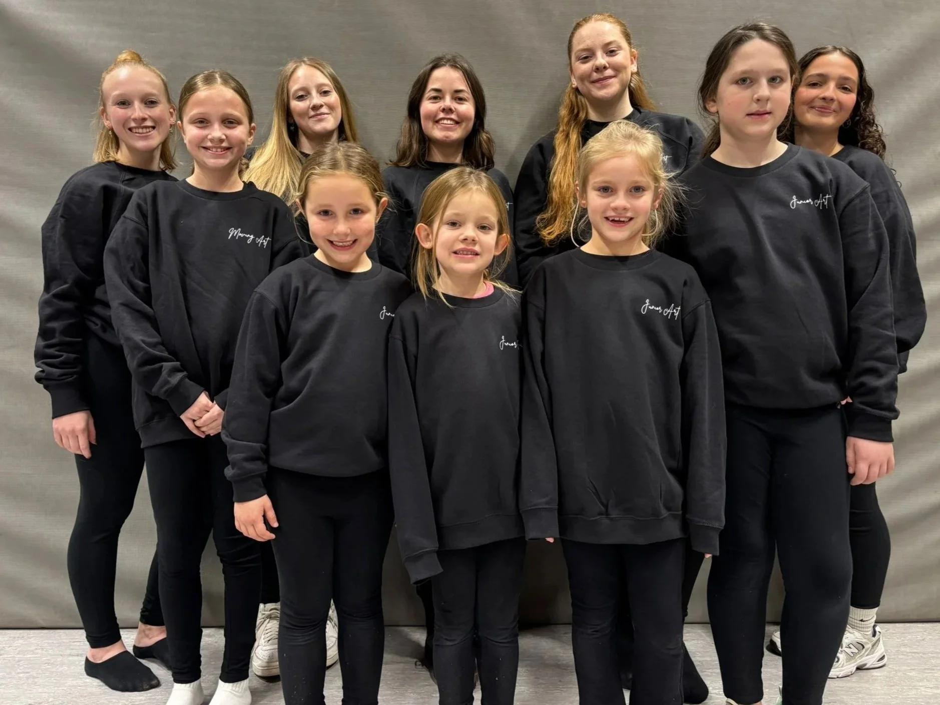 Tien jonge meisjes staan samen, gekleed in zwarte sweatshirts met de tekst 'Junior Art' erop, op een neutrale achtergrond, glimlachend naar de camera.