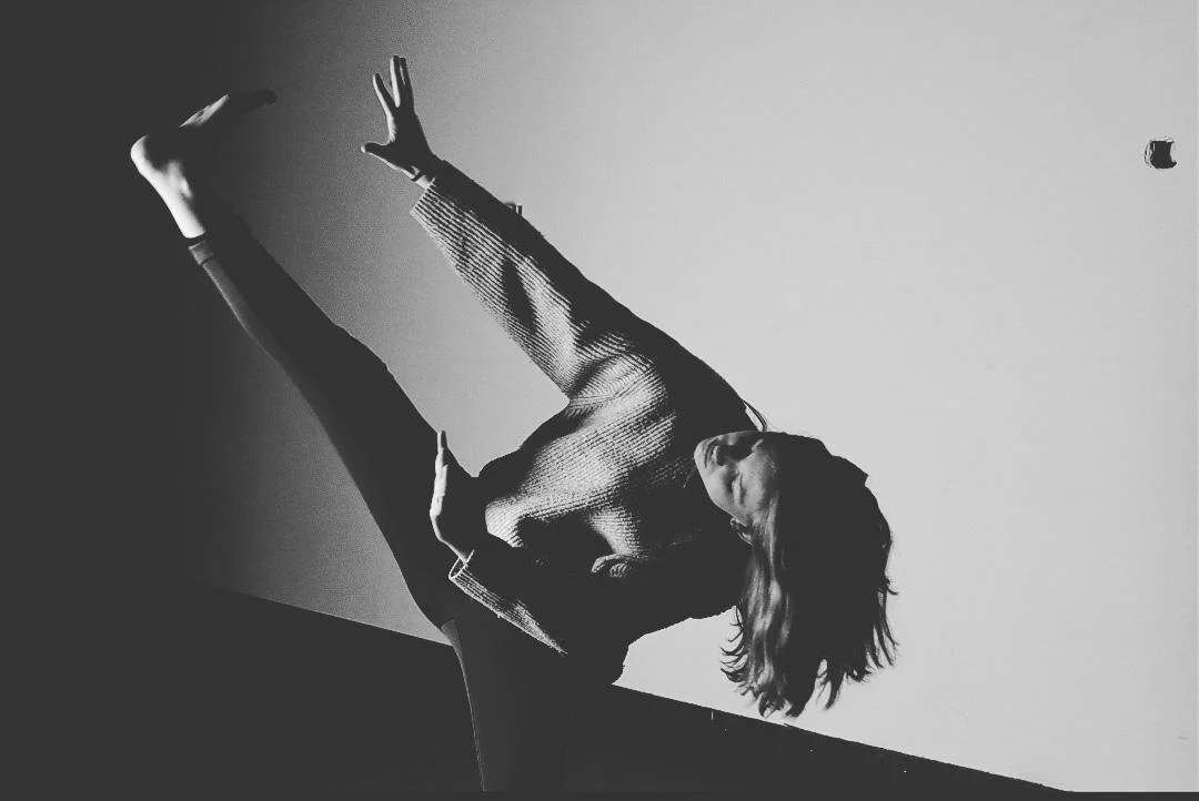 Zwart-wit foto van een vrouw die een danspositie aanneemt, op een diagonale lijn tegen een lege muur.