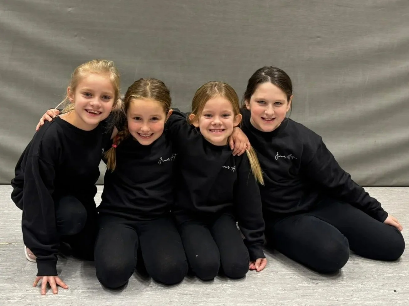 Vijf jonge meisjes zitten op de vloer, gekleed in zwarte sportkleding, en glimlachen naar de camera.