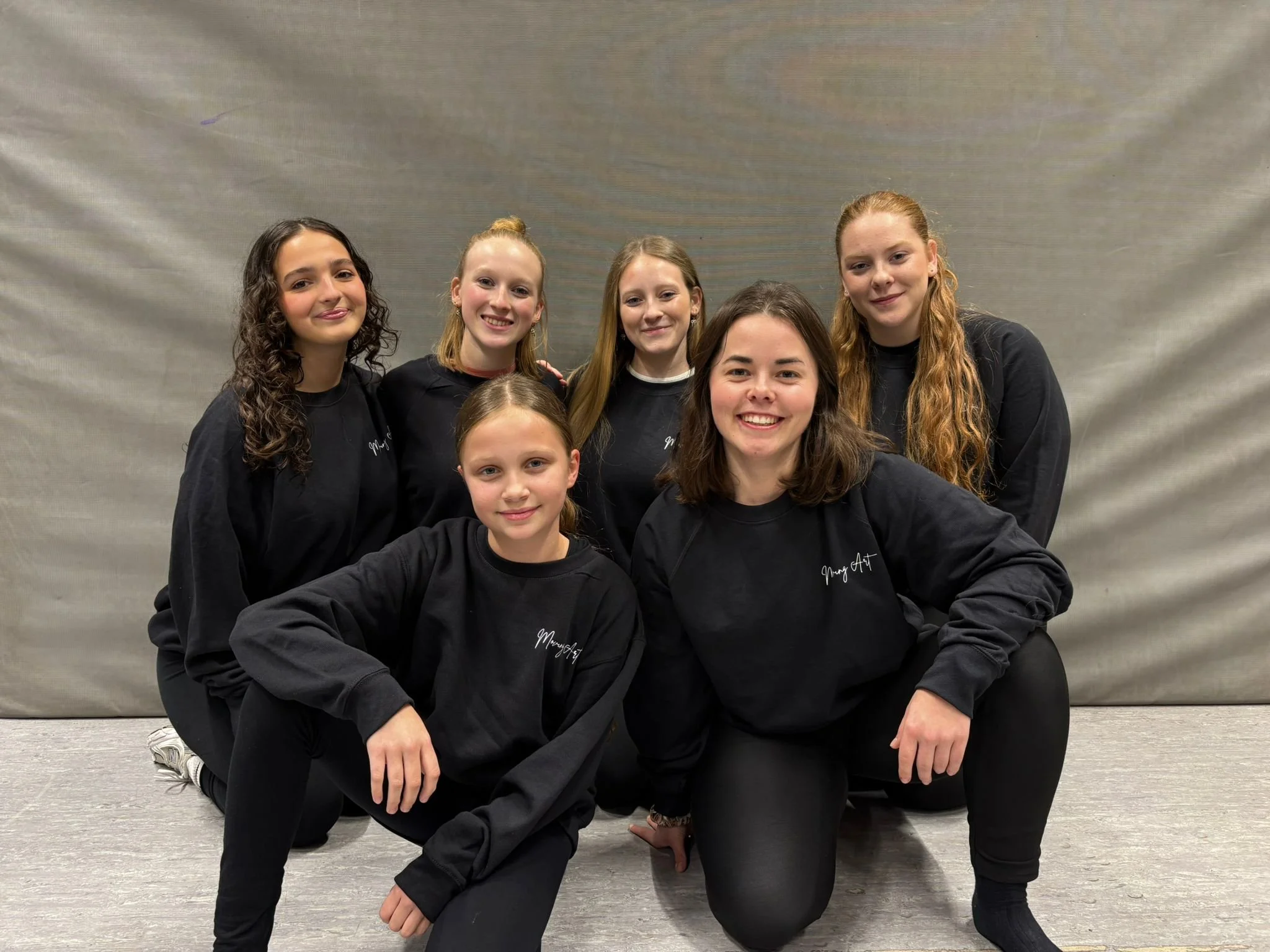 Zeven jonge vrouwen in zwarte sweatshirts met witte tekst, poseren voor een effen grijze achtergrond.