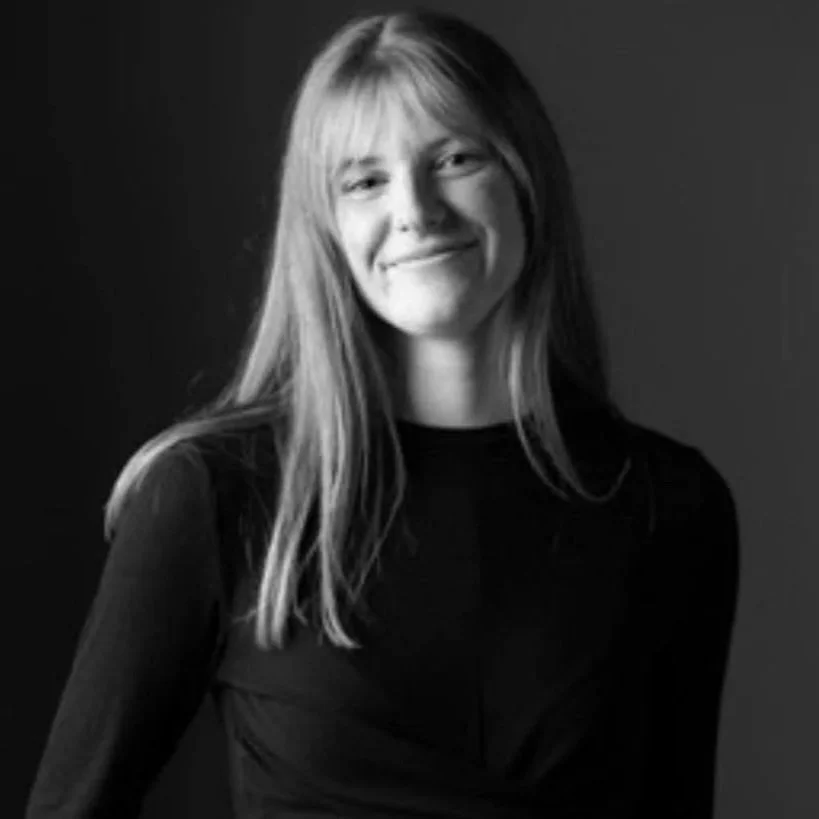 Zwarte en witte portretfoto van een glimlachende vrouw met lang haar, gekleed in een zwarte top, tegen een donkere achtergrond.