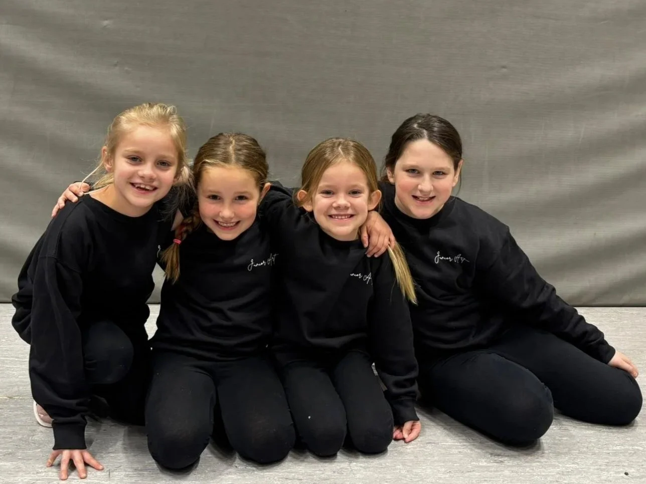 Vier jonge meisjes in zwarte sportkleding zitten knielend en lachen samen voor een grijze achtergrond.