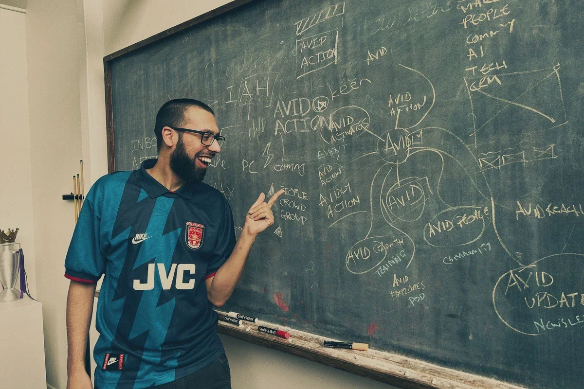 A man with glasses and a beard, smiling and pointing at a classroom blackboard filled with notes and diagrams about 'AVID' and 'AVID action'.
