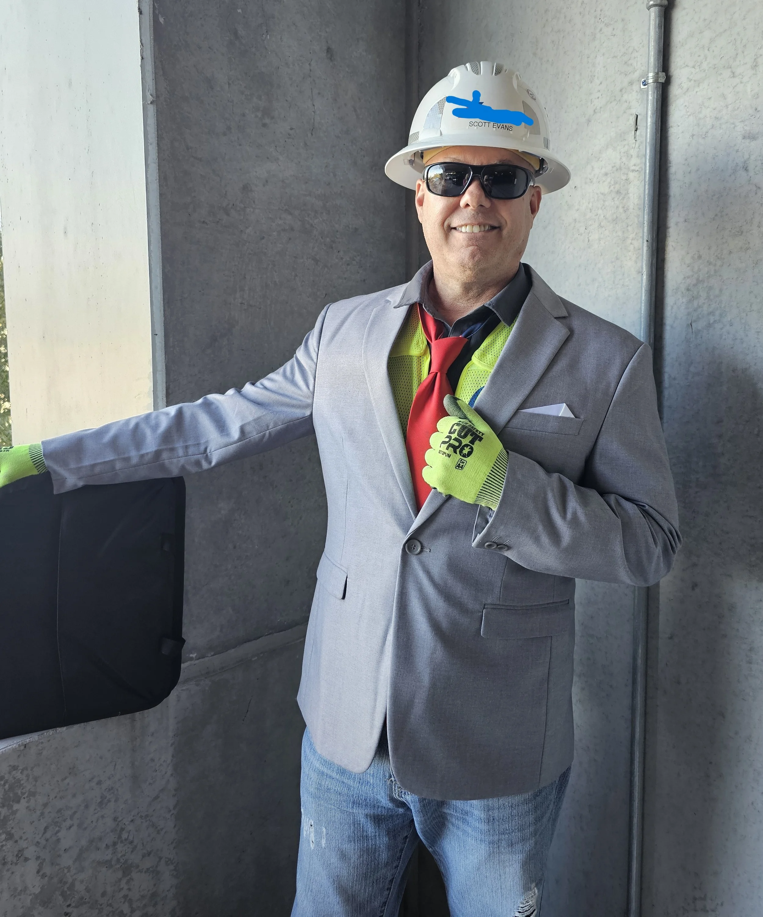 Scott Evans wearing suit, tie, hard hat and safety vest, smiling and pointing during jobsite visit.