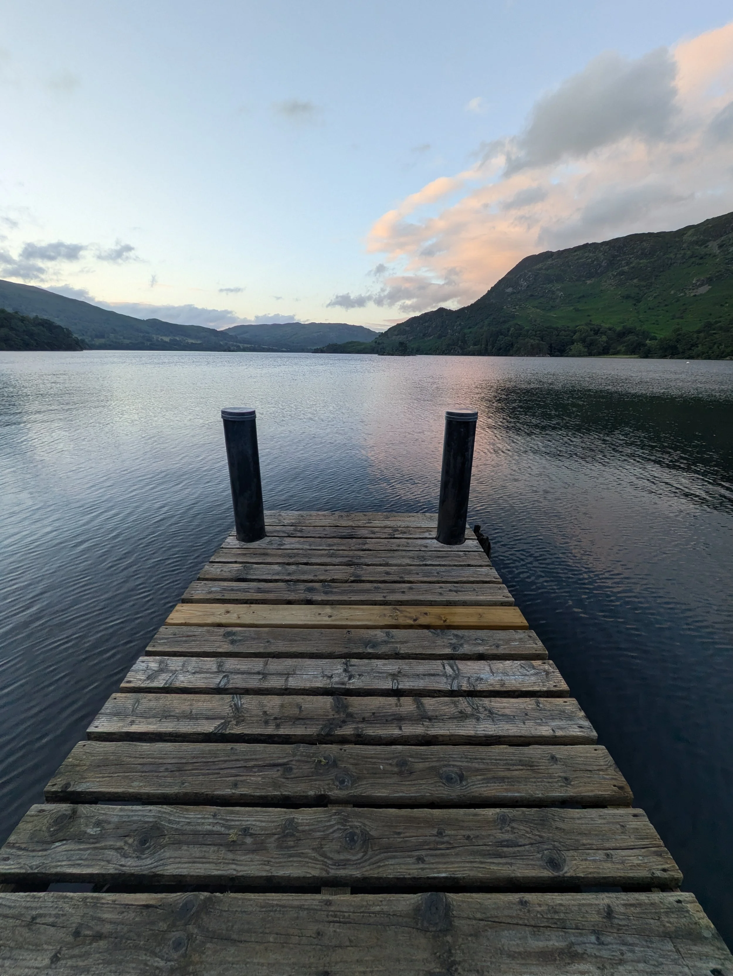 A wooden dock extending over a Ullswater, with hills covered in greenery and a partly cloudy sky in the background.