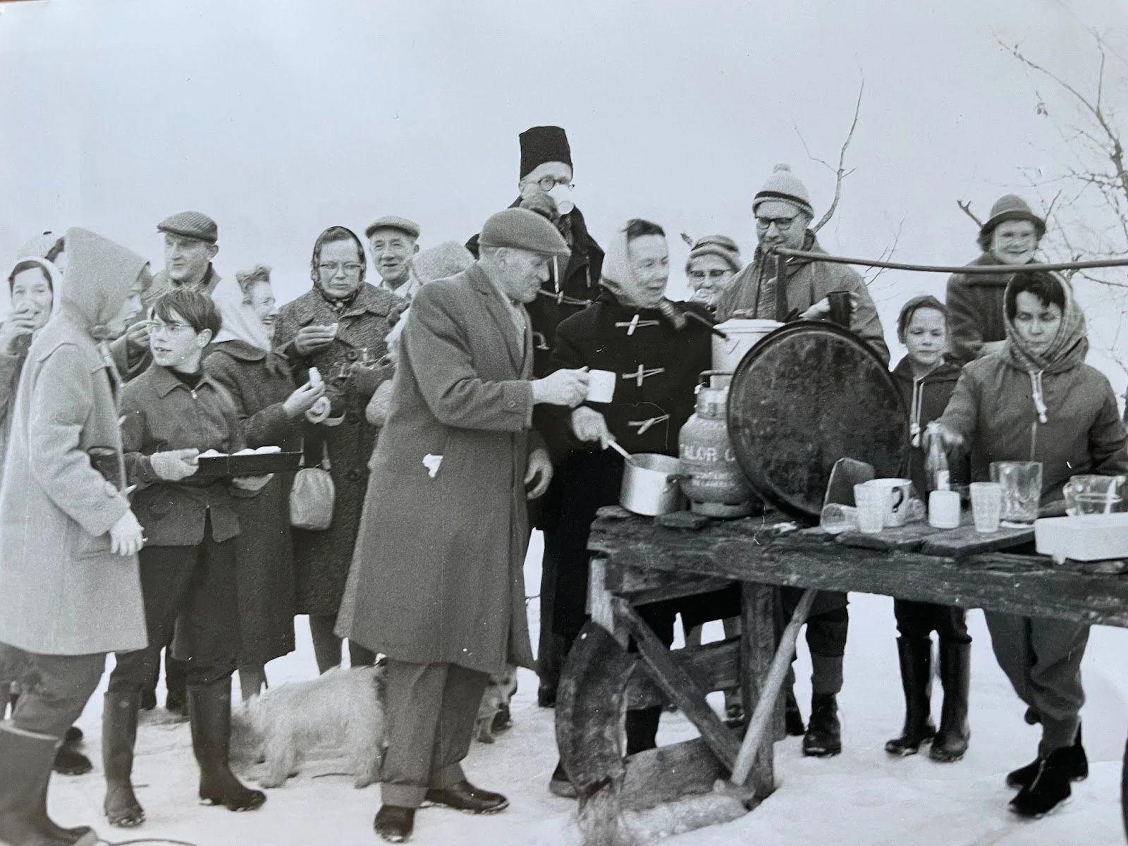 Black and white photo of a group of people gathered around a food stall outdoors on snowy ground, serving hot drinks during the Big Freeze