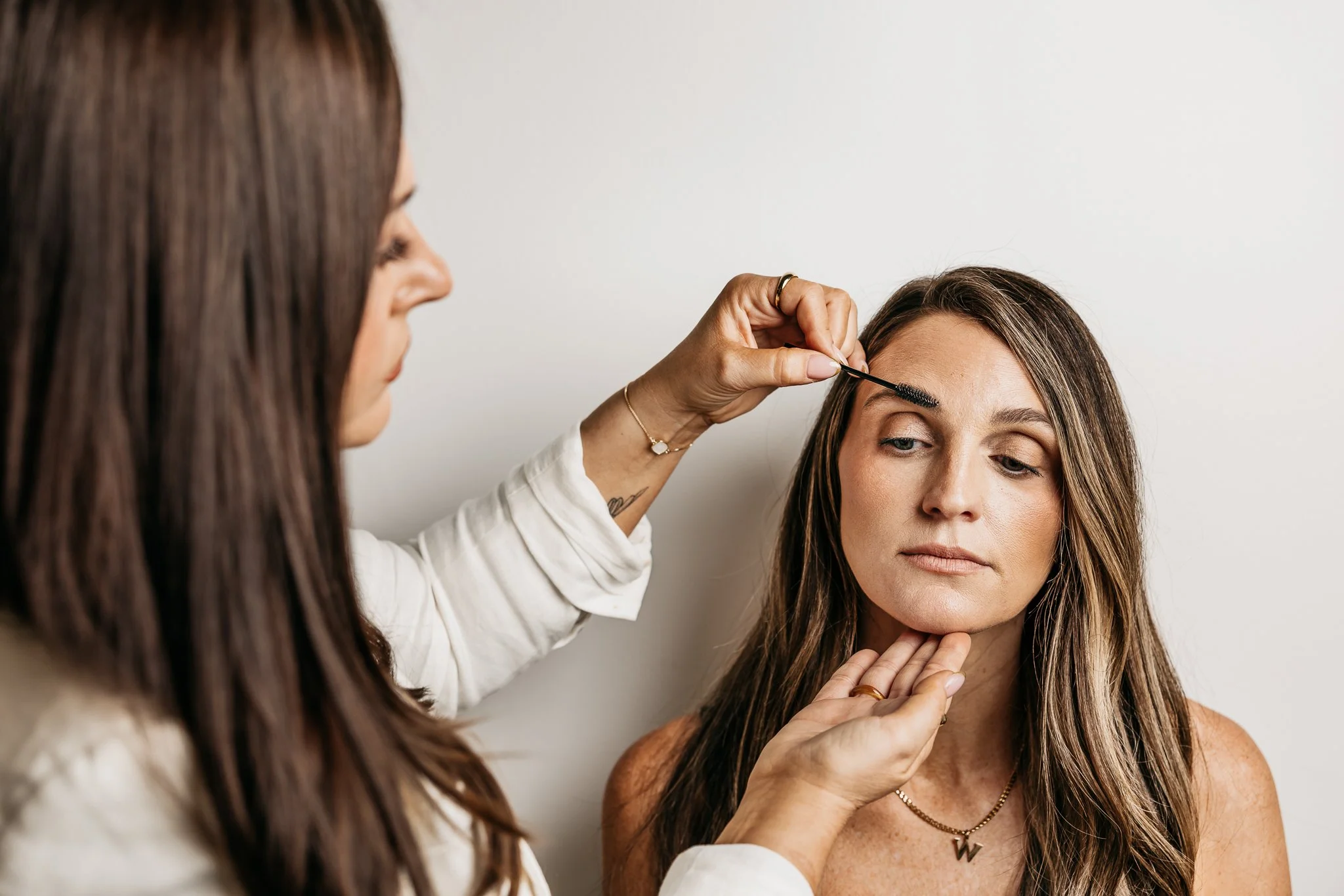 A woman with long brown hair is having her eyebrows shaped by a beautician with long brown hair, wearing a white shirt, against a white wall.