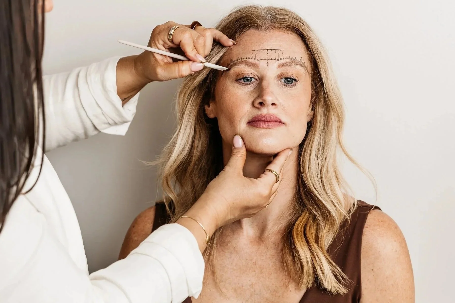 A woman is getting an aesthetic procedure, with a mark and guide on her forehead, while a practitioner measures her face with her hand under her chin.